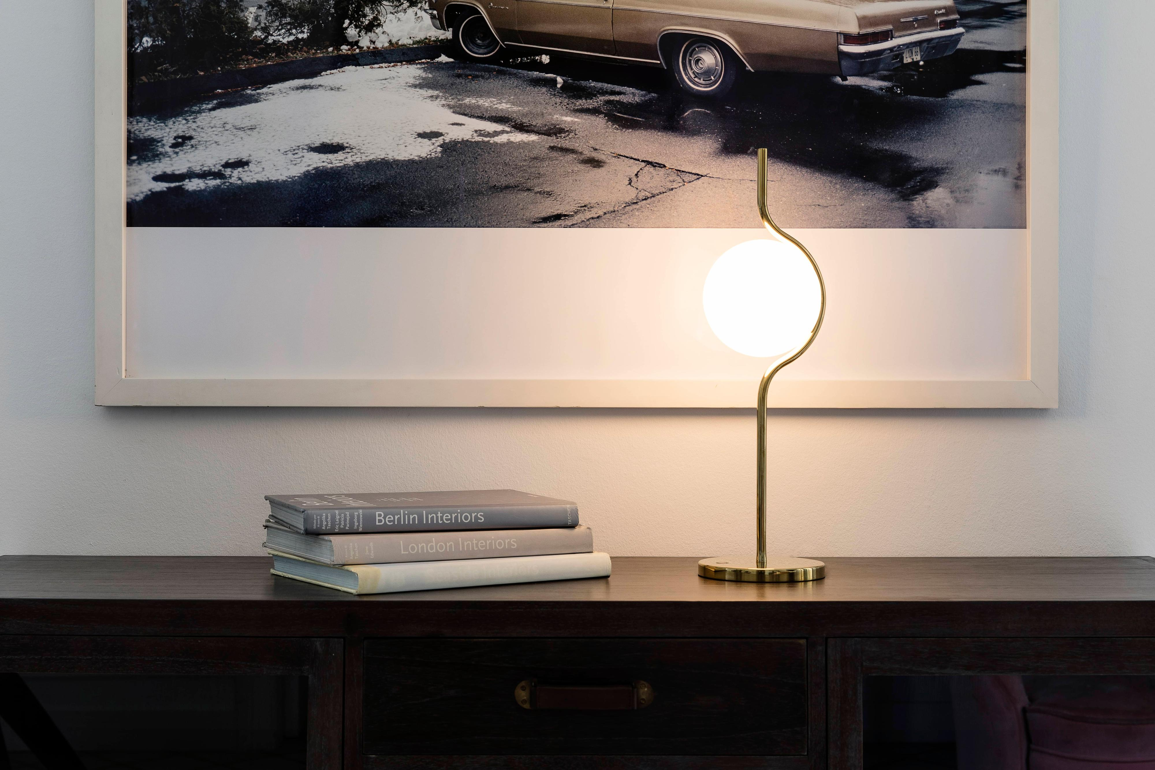 Lampada dorata con globo bianco, libri e superficie in legno scuro con dipinto dietro.