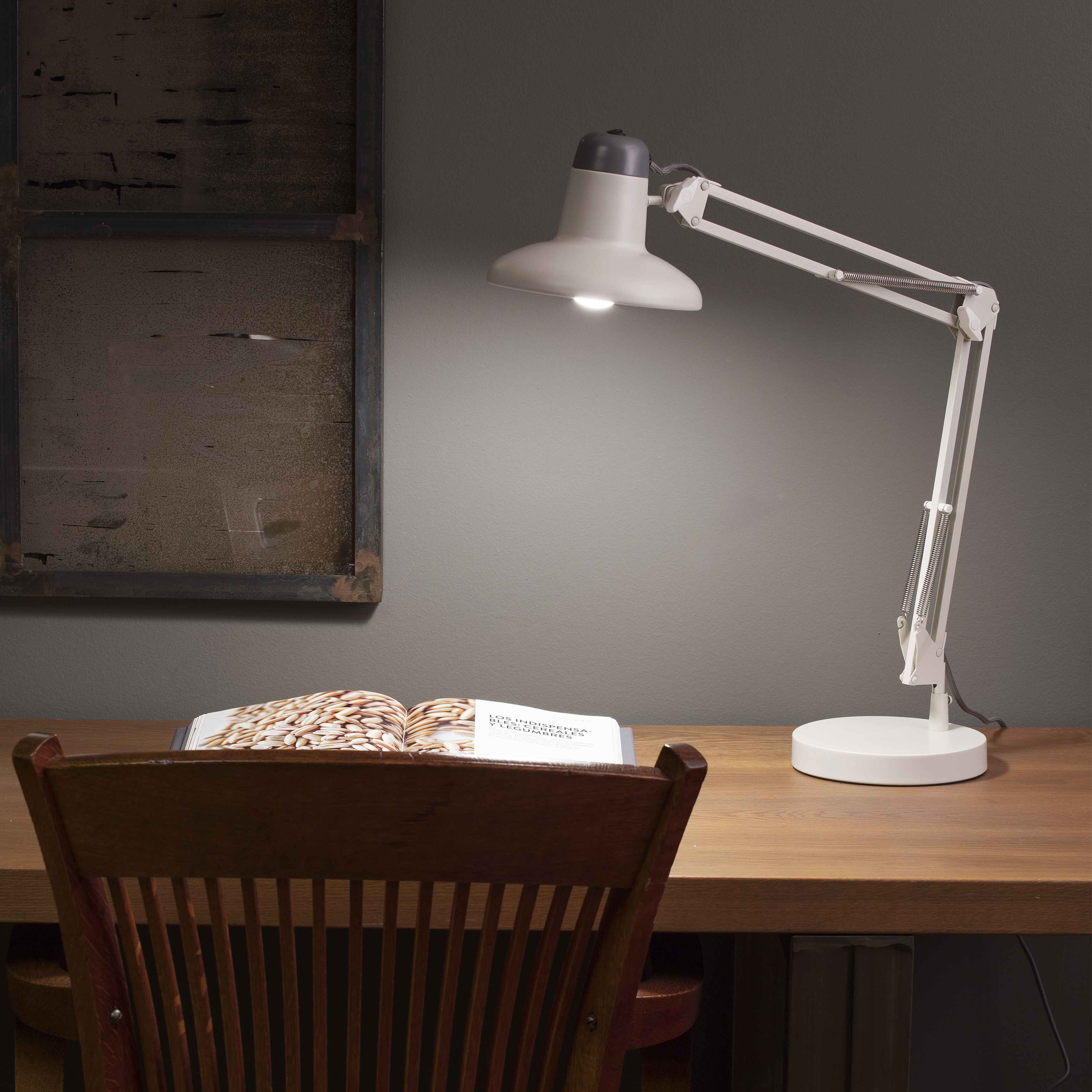 Weiße Schreibtischlampe mit Buch auf Holztisch. Fenster und Stuhl im Hintergrund.