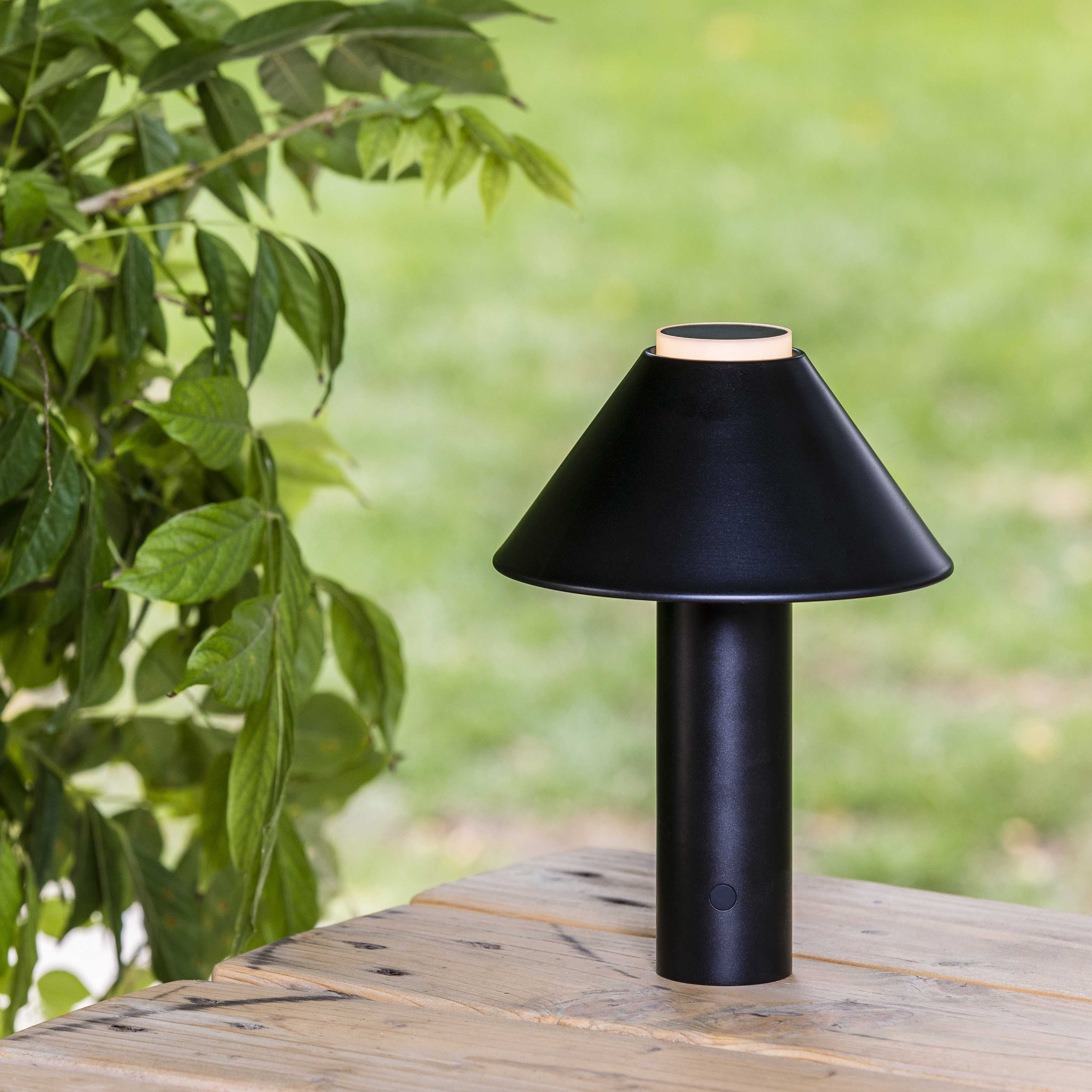 Lampada da tavolo nera con paralume conico, su un tavolo di legno. Fogliame verde sullo sfondo.