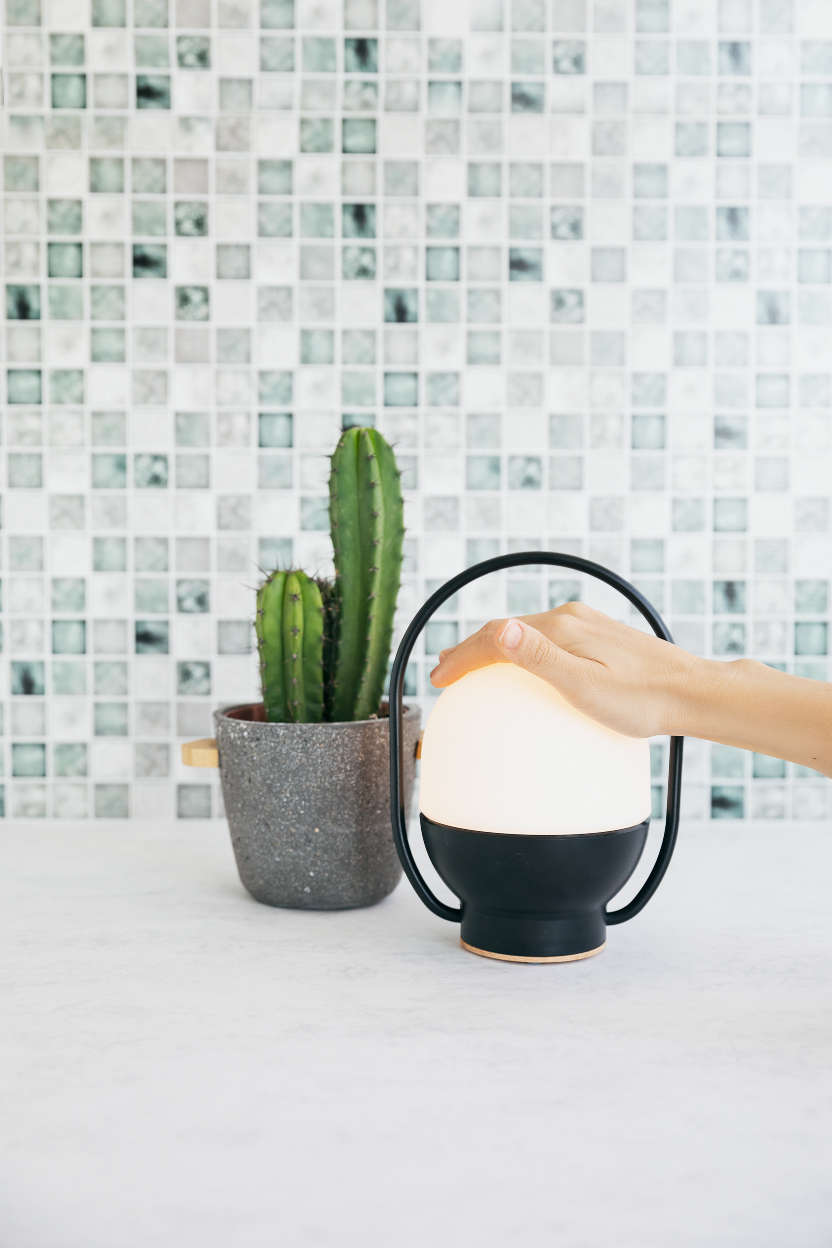 Una mano tiene in mano una lampada bianca brillante con un manico nero e un cactus verde accanto.