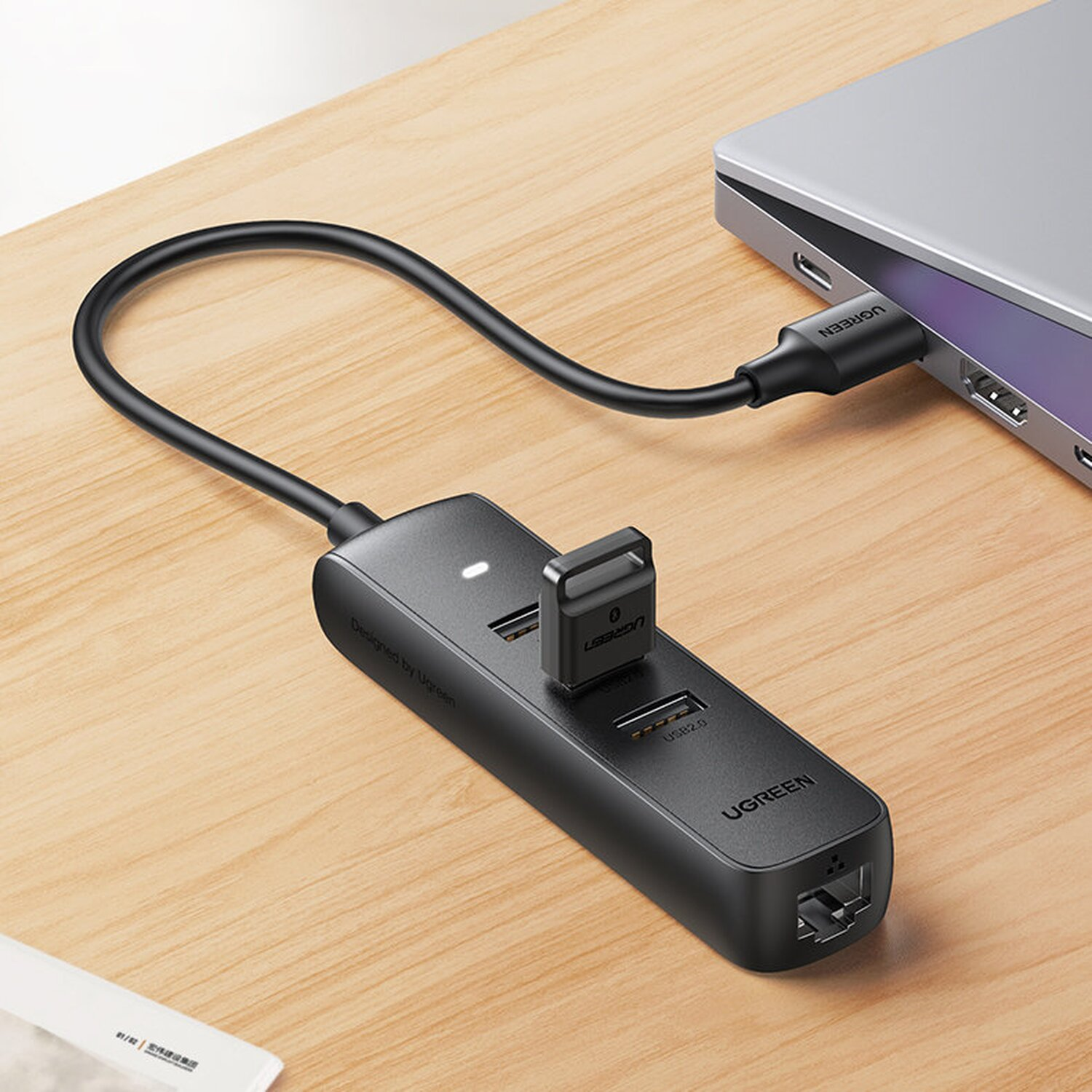 Schwarzer UGREEN USB-Hub mit angeschlossenem USB-C-Kabel, einem USB-Stick und einem Laptop auf einem Holzschreibtisch.