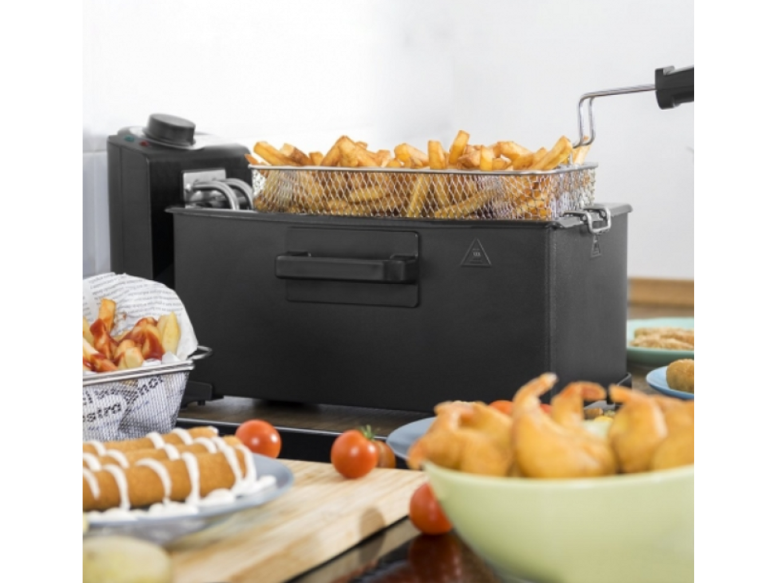 Frytownica z frytkami w koszu, otoczona innymi smażonymi potrawami i pomidorami.