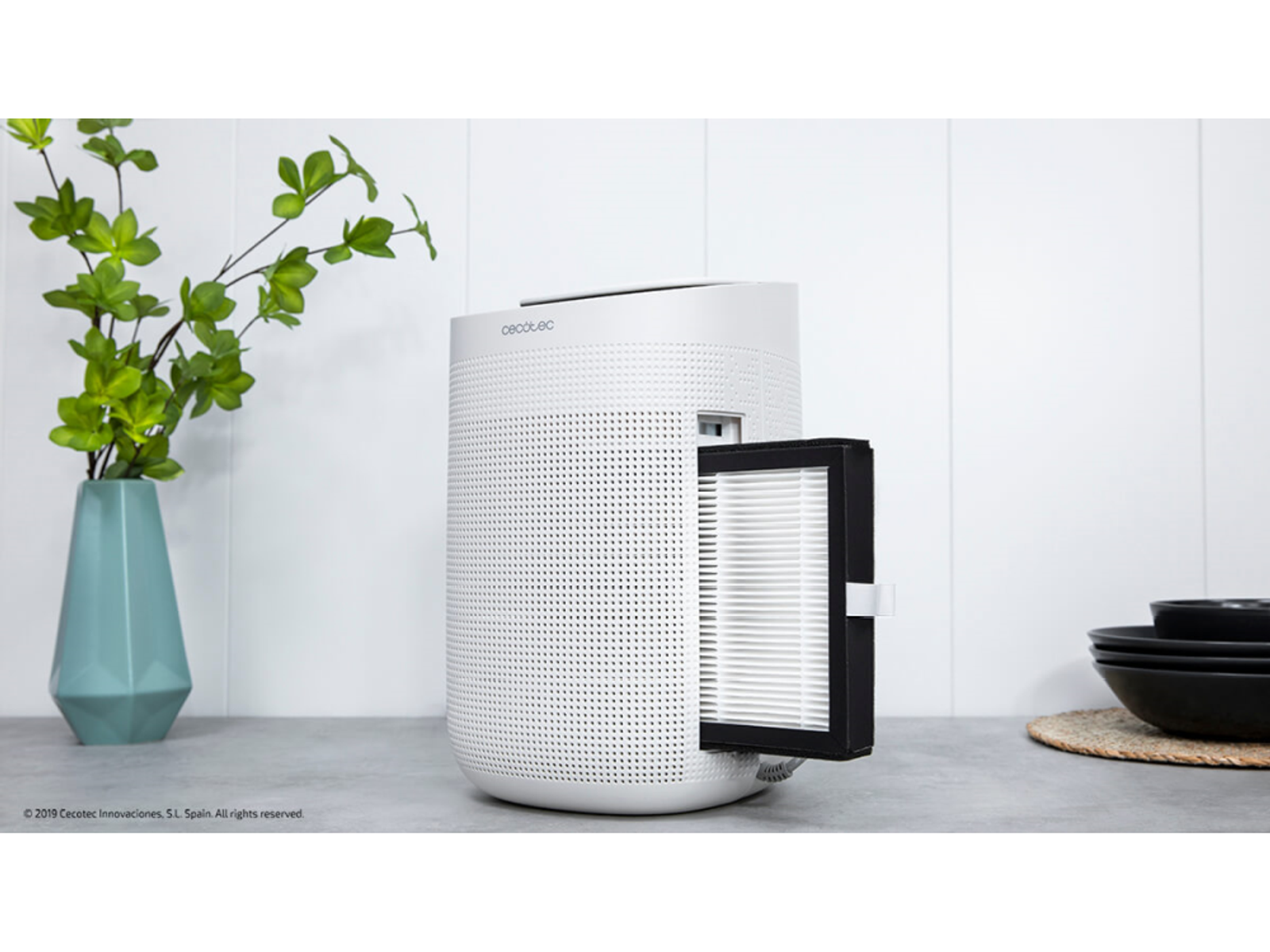 Biały oczyszczacz powietrza z wyjmowanym filtrem, z wazonem zielonych roślin w tle.