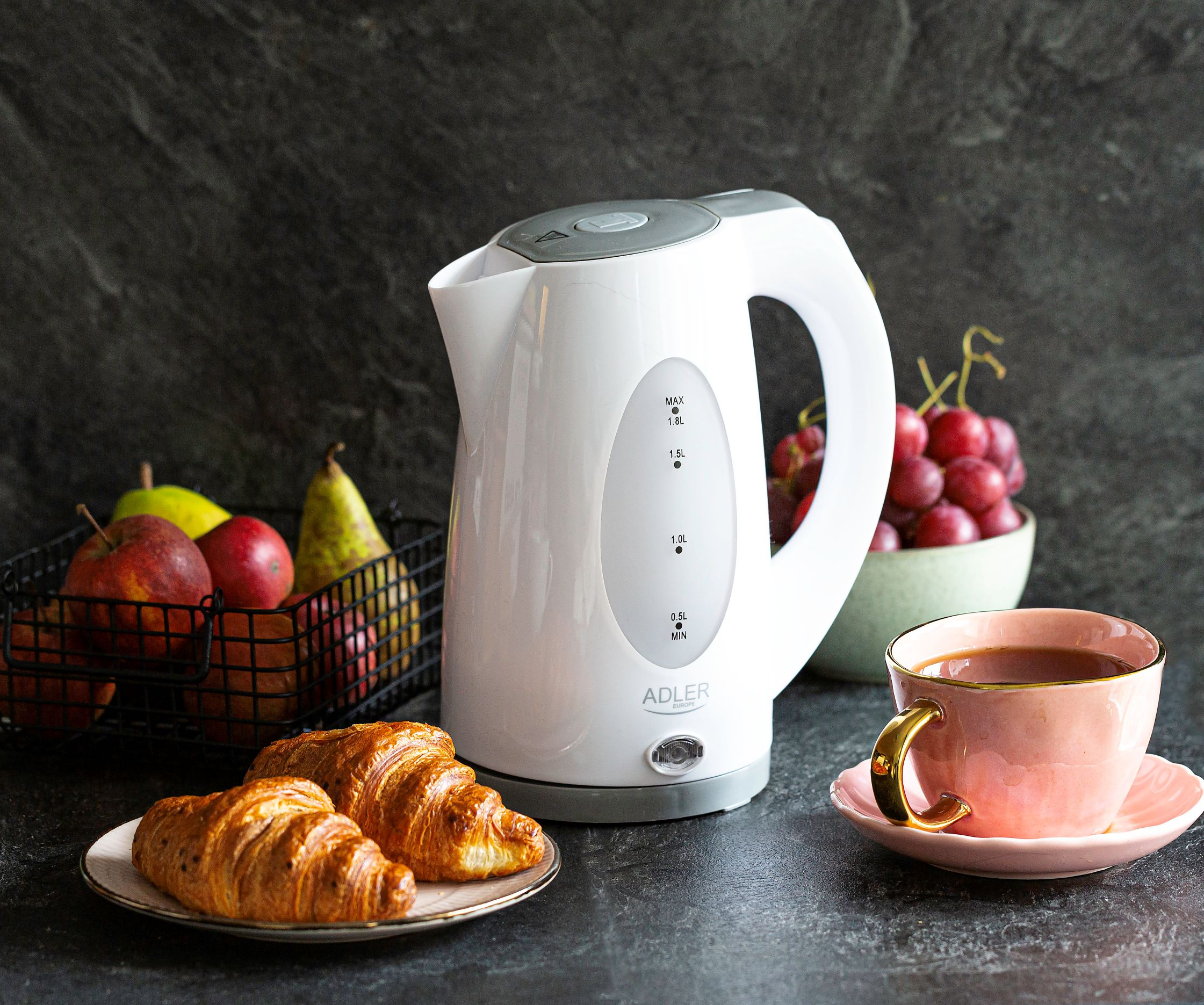 Weißer Wasserkocher mit Obst, Croissants und Teetasse auf einem Tisch. Dunkler Hintergrund.