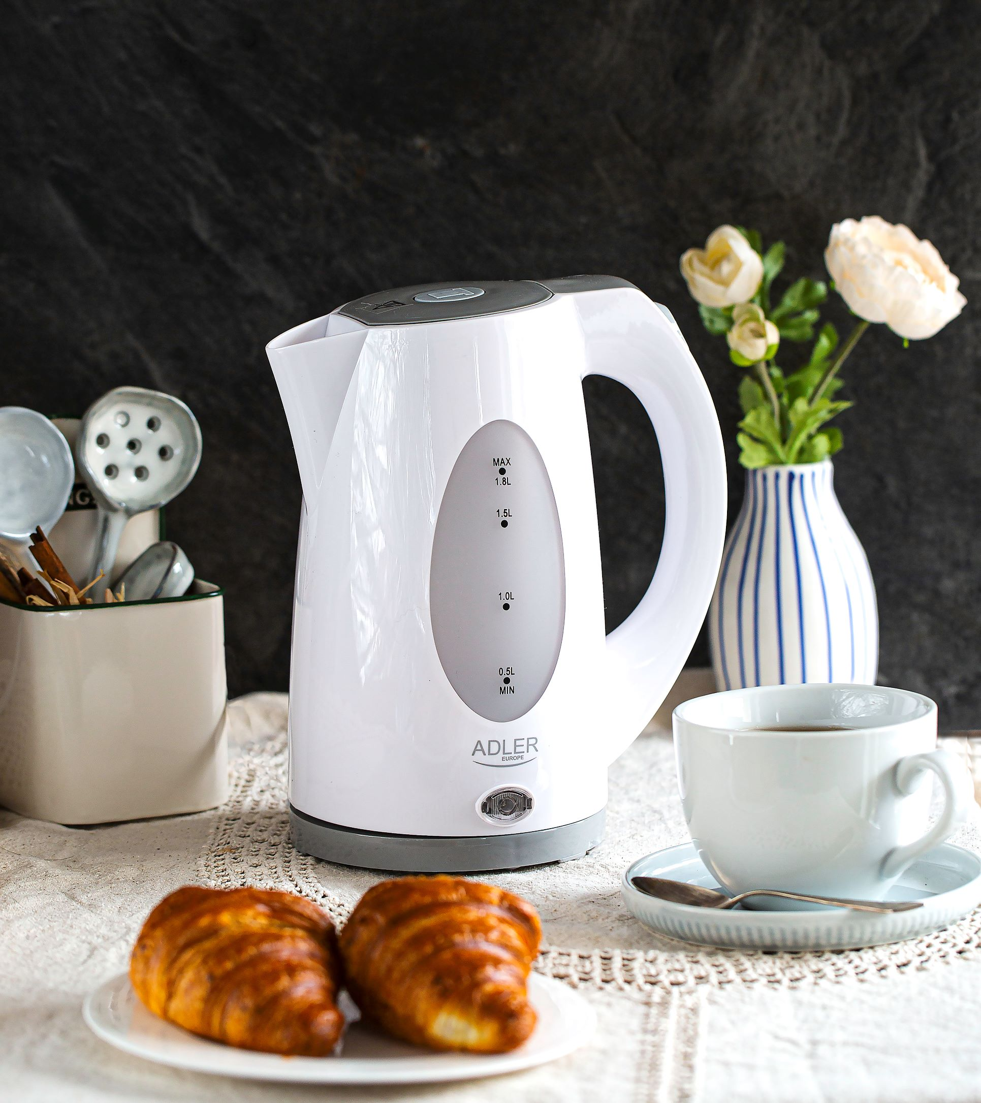 Weißer Wasserkocher mit Croissants auf einem Teller, einer Tasse Kaffee und Blumen.