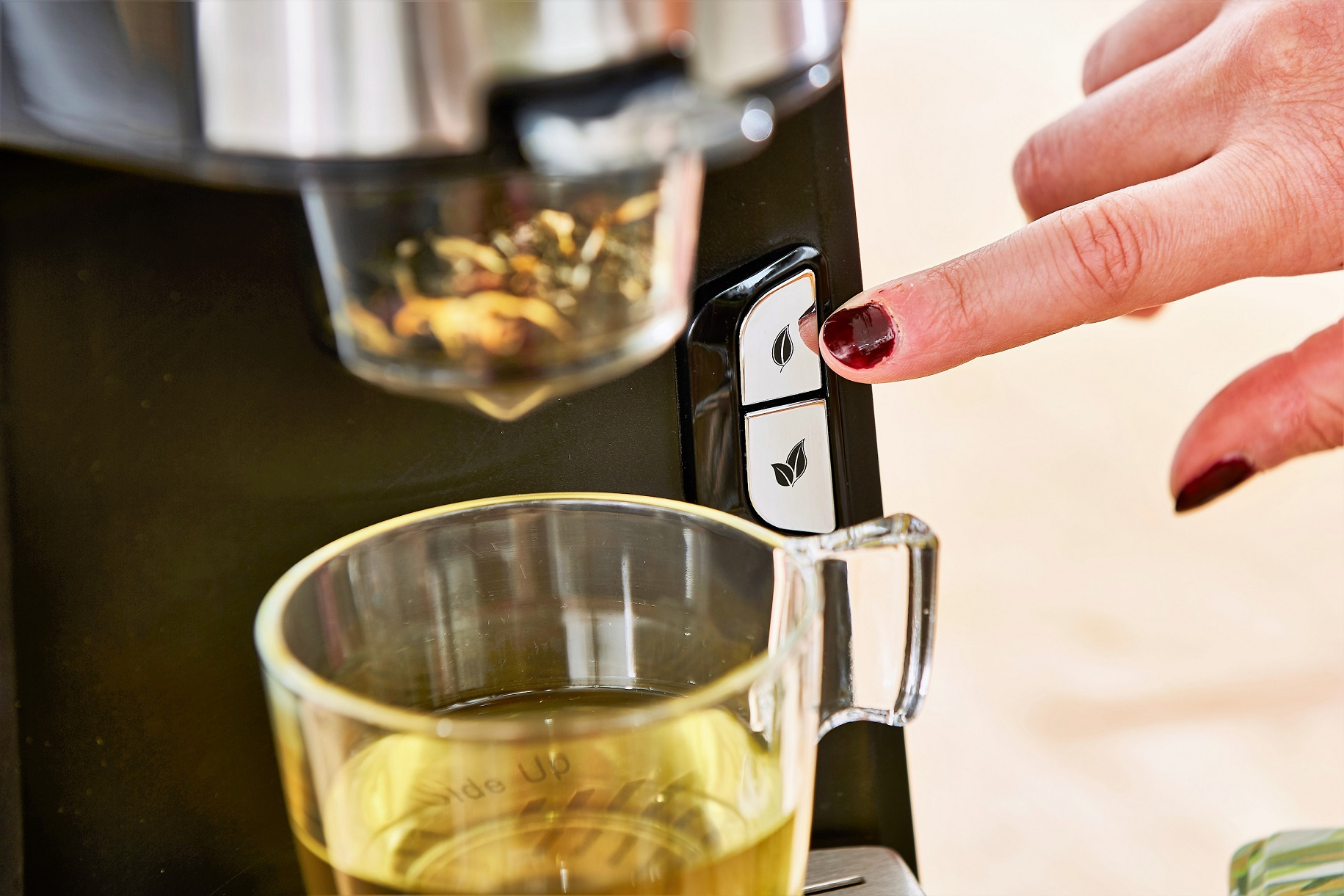 Die Hand einer Frau drückt eine Taste an einer schwarzen Teemaschine. Ein Glas enthält grünen Tee.