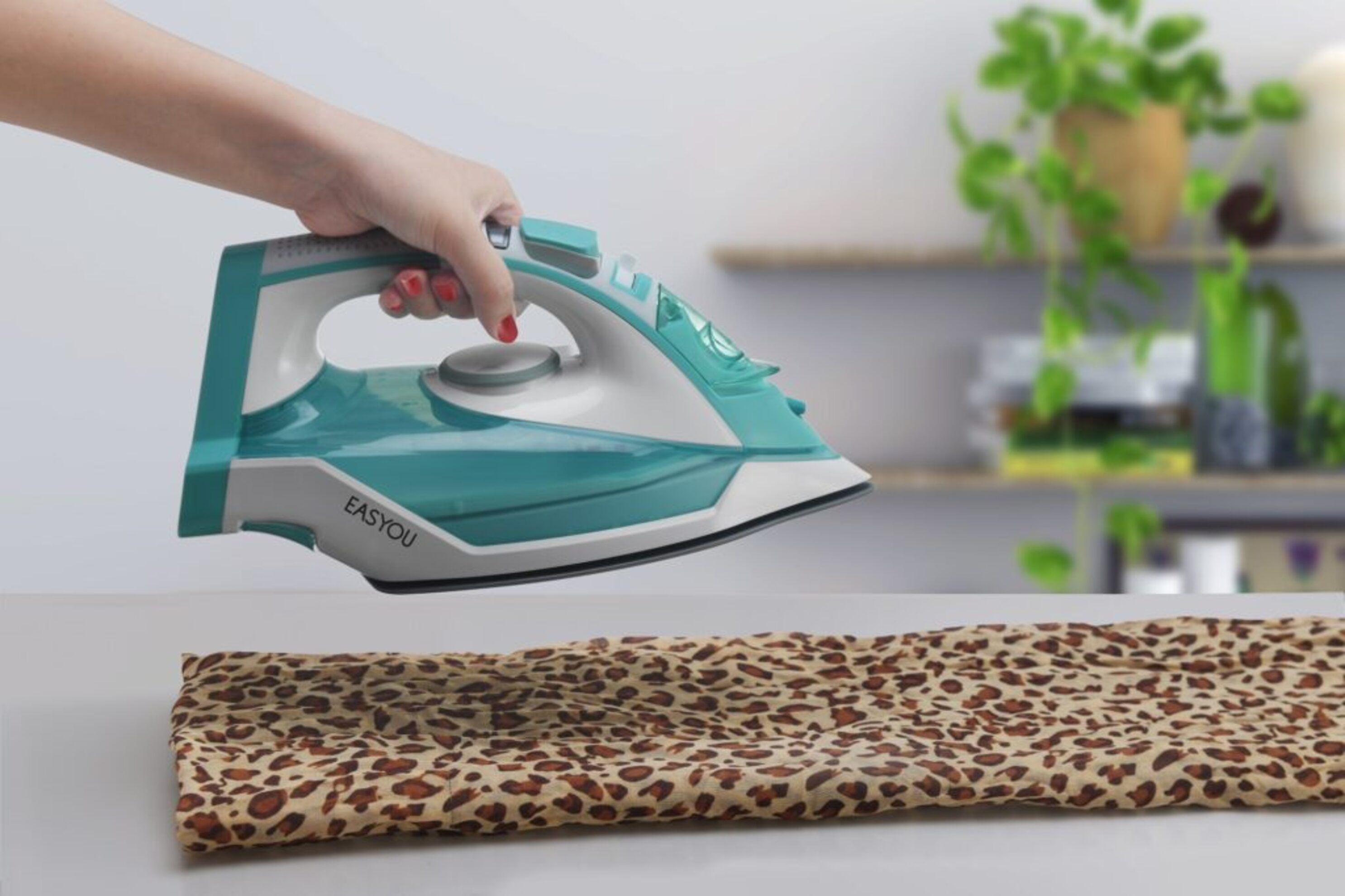 Mano planchando una tela con estampado de leopardo sobre una superficie blanca con plantas.