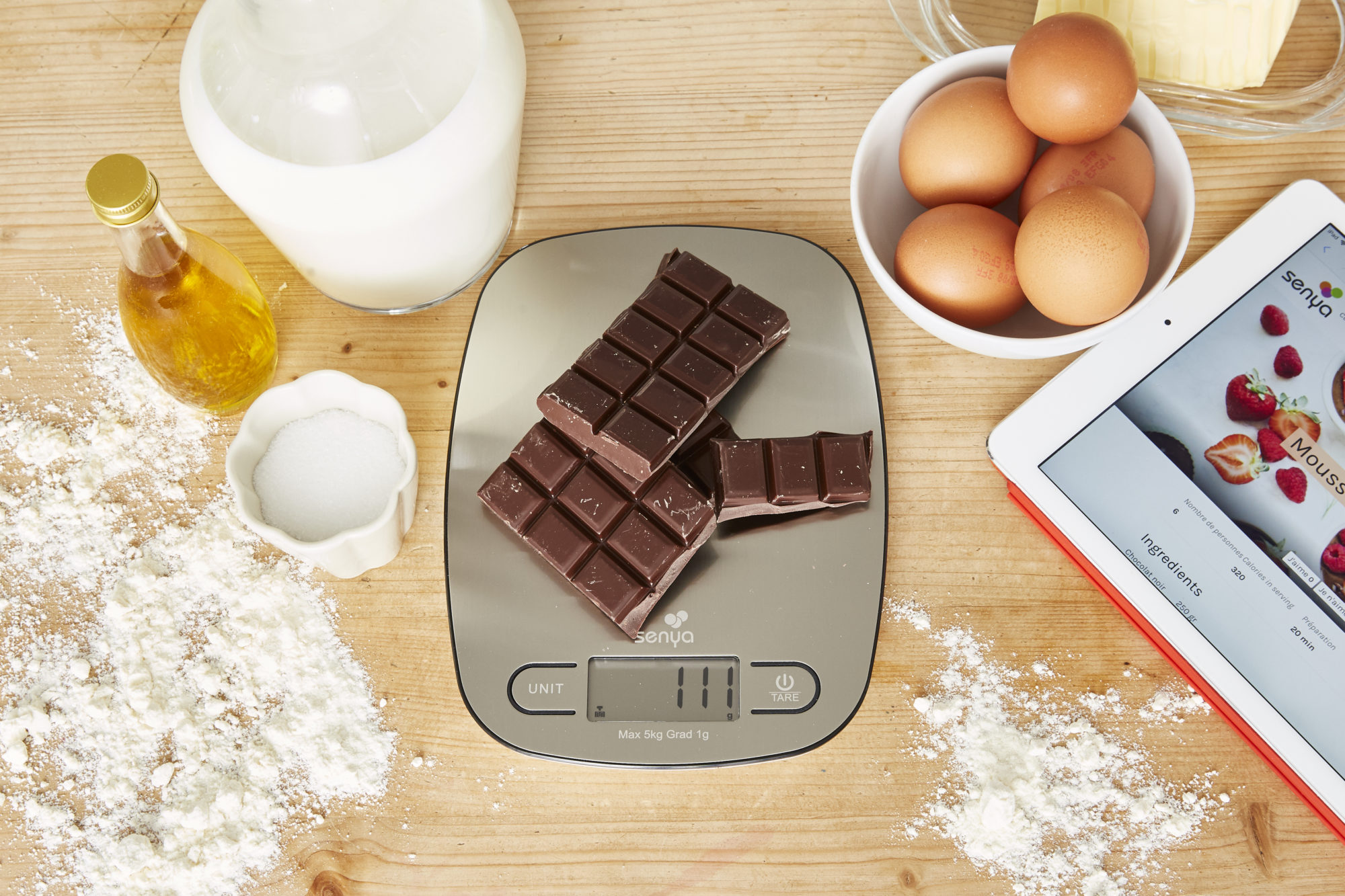 Schokoladenriegel auf einer Waage, umgeben von Eiern, Öl, Zucker und einem Tablet mit Rezept.