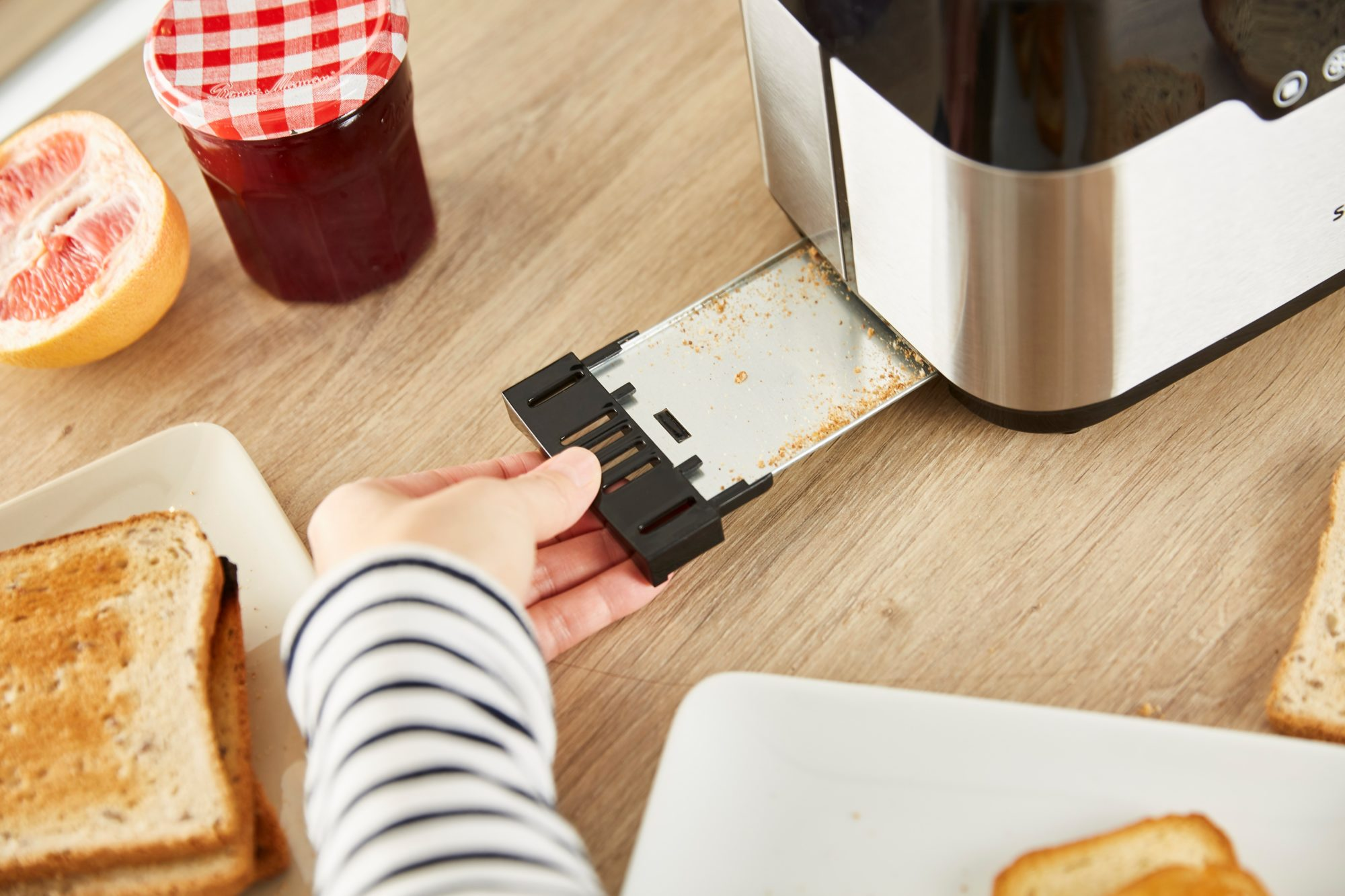 Hand zieht einen Toaster-Rack aus einem silbernen Toaster auf einem Holztisch. Andere Gegenstände sind sichtbar.