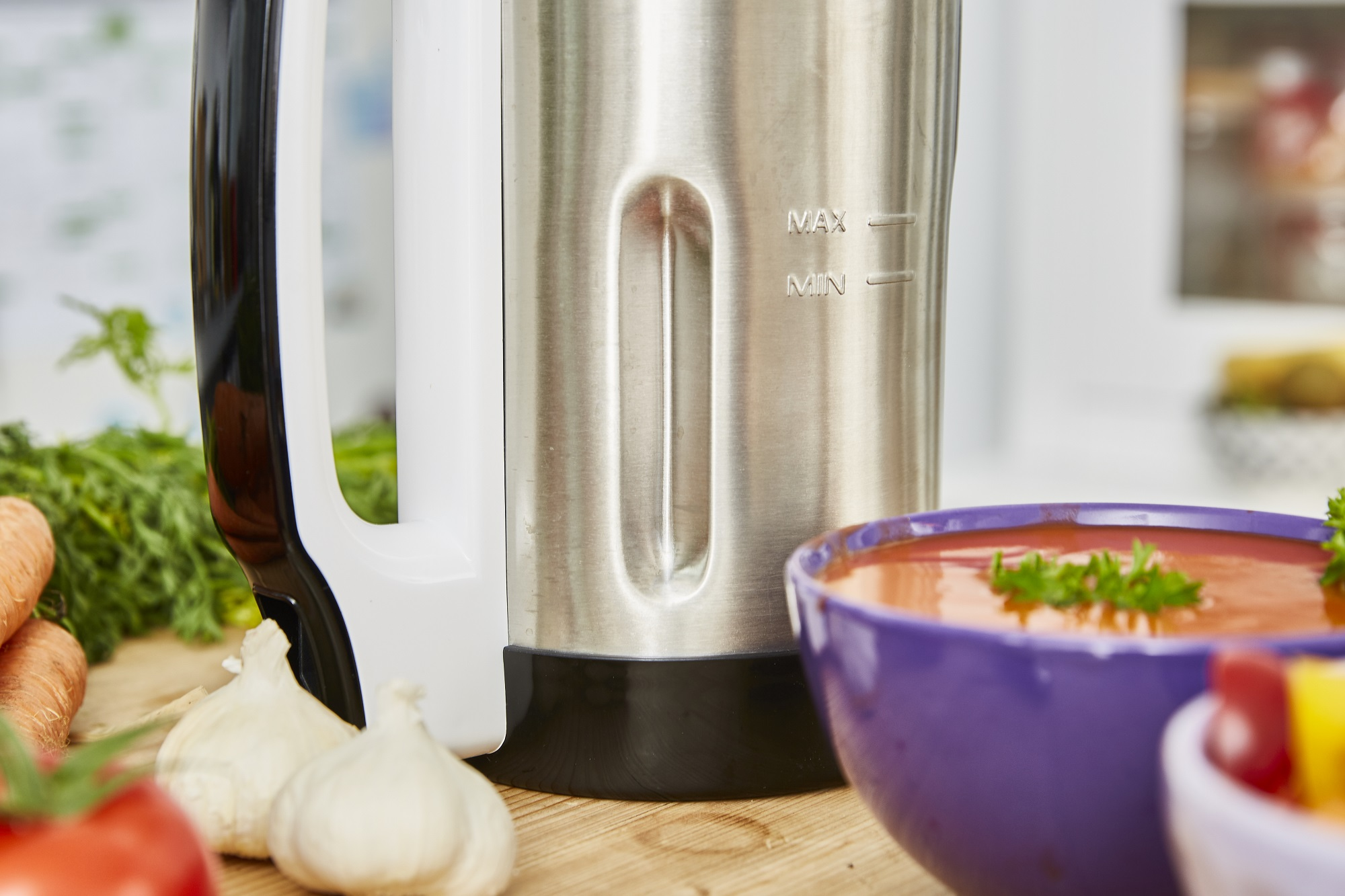 Nahaufnahme eines Edelstahl-Mixers mit Griff und einer Schüssel roter Suppe, mit Gemüse.