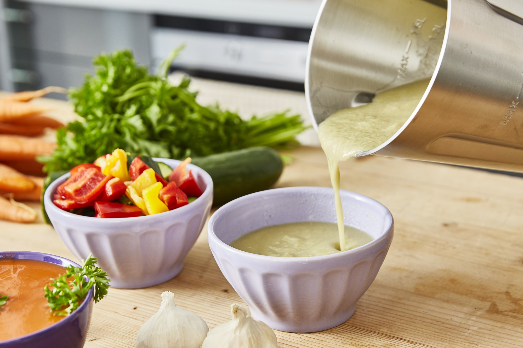 Eine Person gießt Suppe in eine Schüssel. Auf einem Holztisch stehen Schüsseln mit Zutaten und Suppe.
