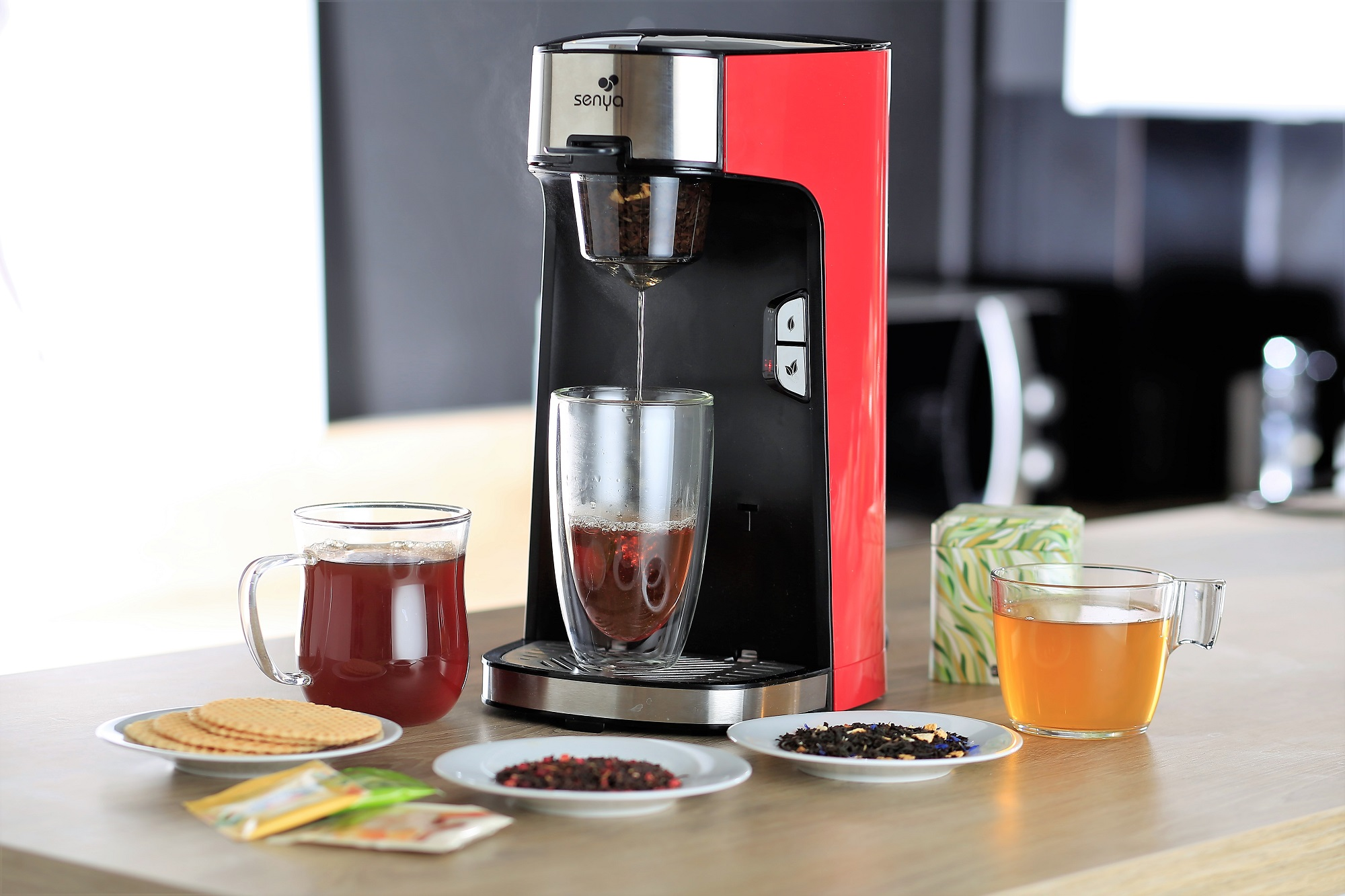 Teemaschine mit Glas. Tees, Waffeln und Teetassen auf einem Tisch.