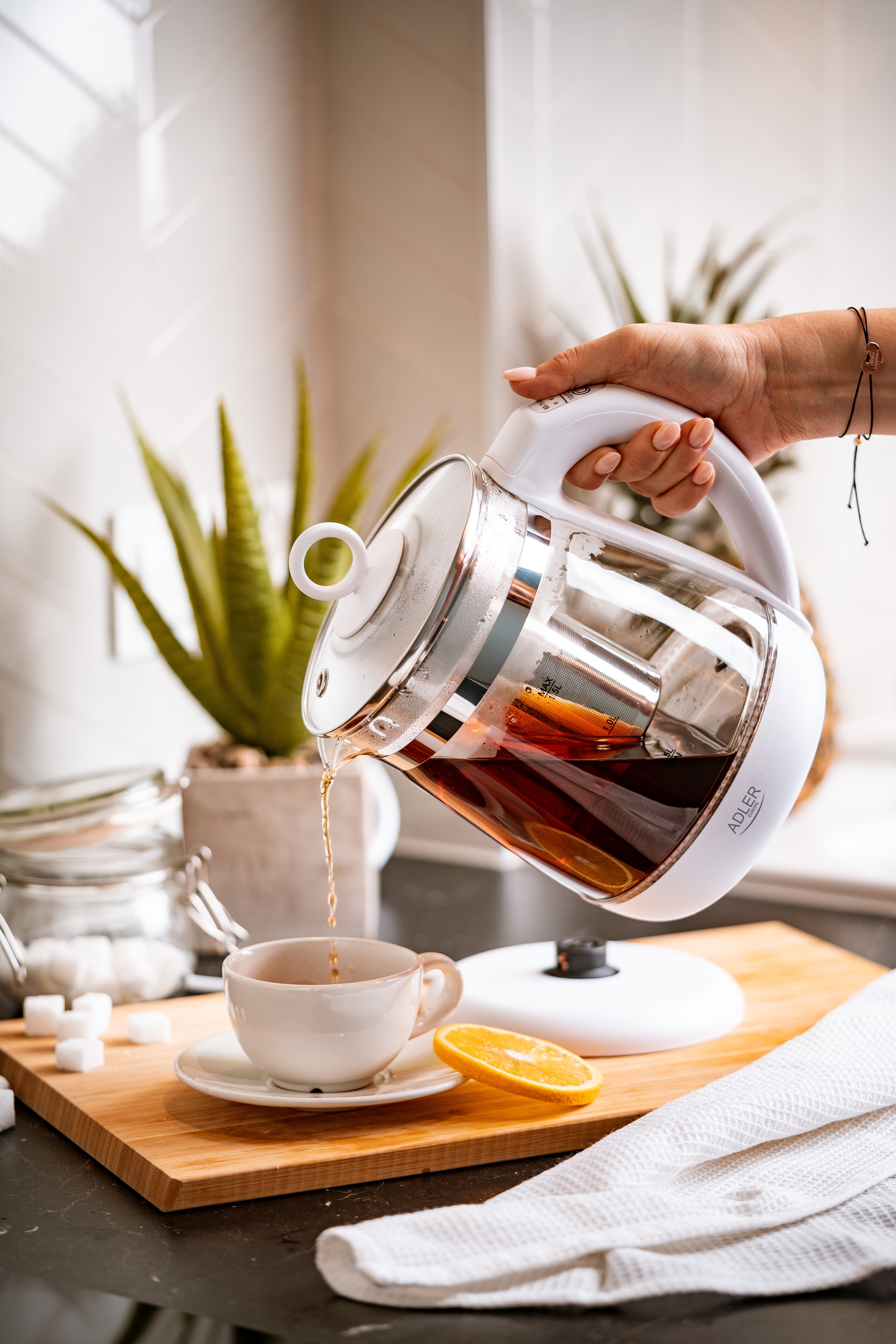 Person gießt Tee aus einem Glas-Teekessel in eine Tasse auf einem Holzbrett.