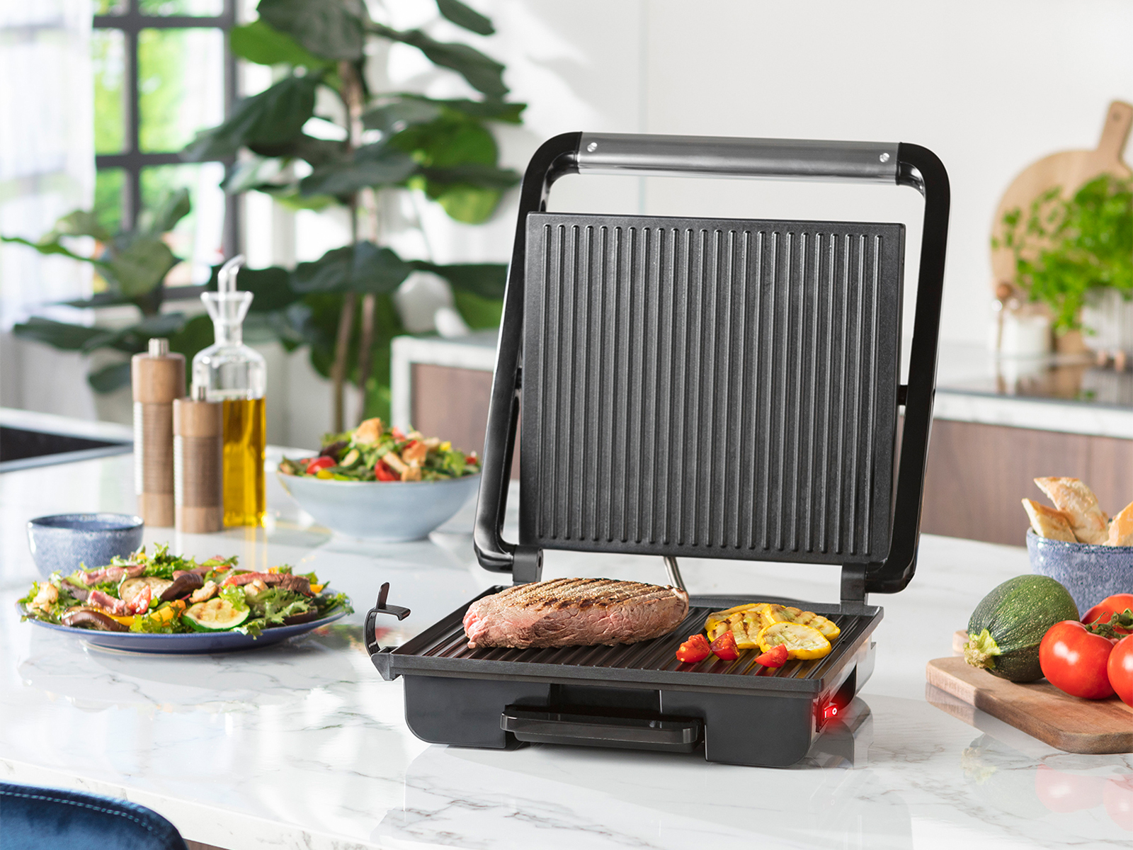 Ein schwarzer Grill mit Essen, darunter ein Steak und Gemüse. Im Hintergrund steht eine Ölflasche.