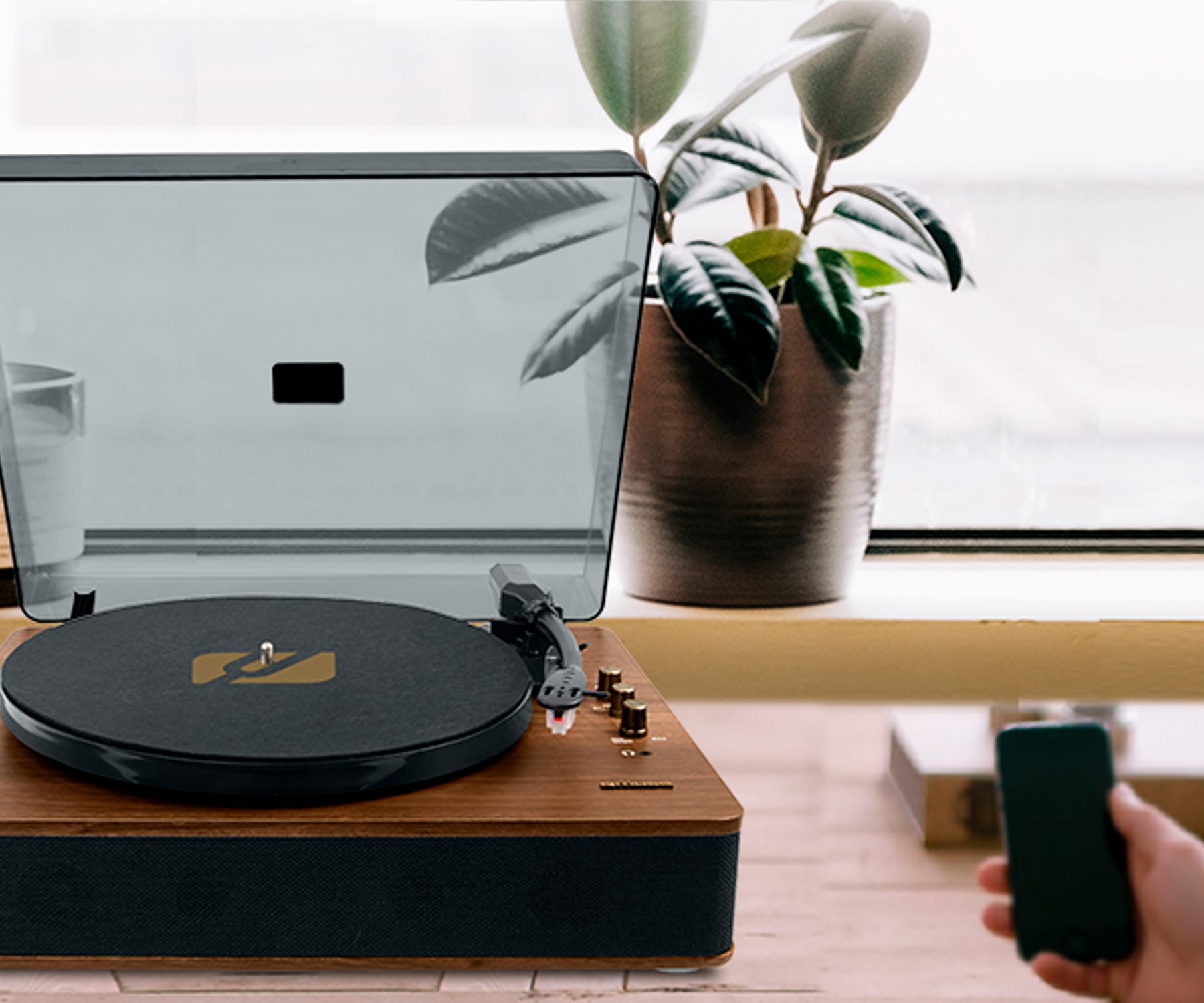 Un tocadiscos de madera con un disco negro, junto a una planta y una persona sosteniendo un teléfono.