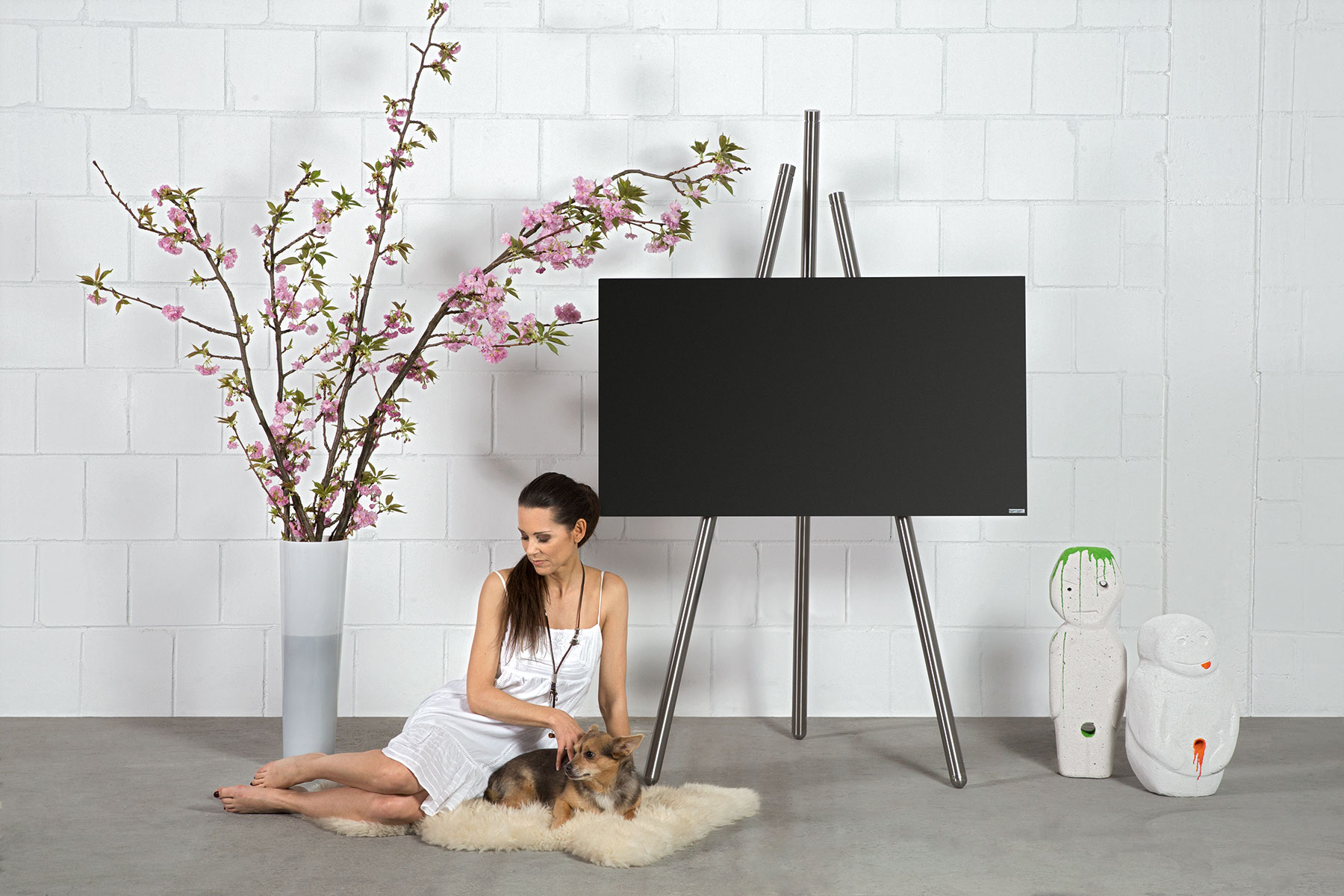 Frau und Hund neben Fernseher auf Ständer mit rosa Blumen und weißer Ziegelwand.