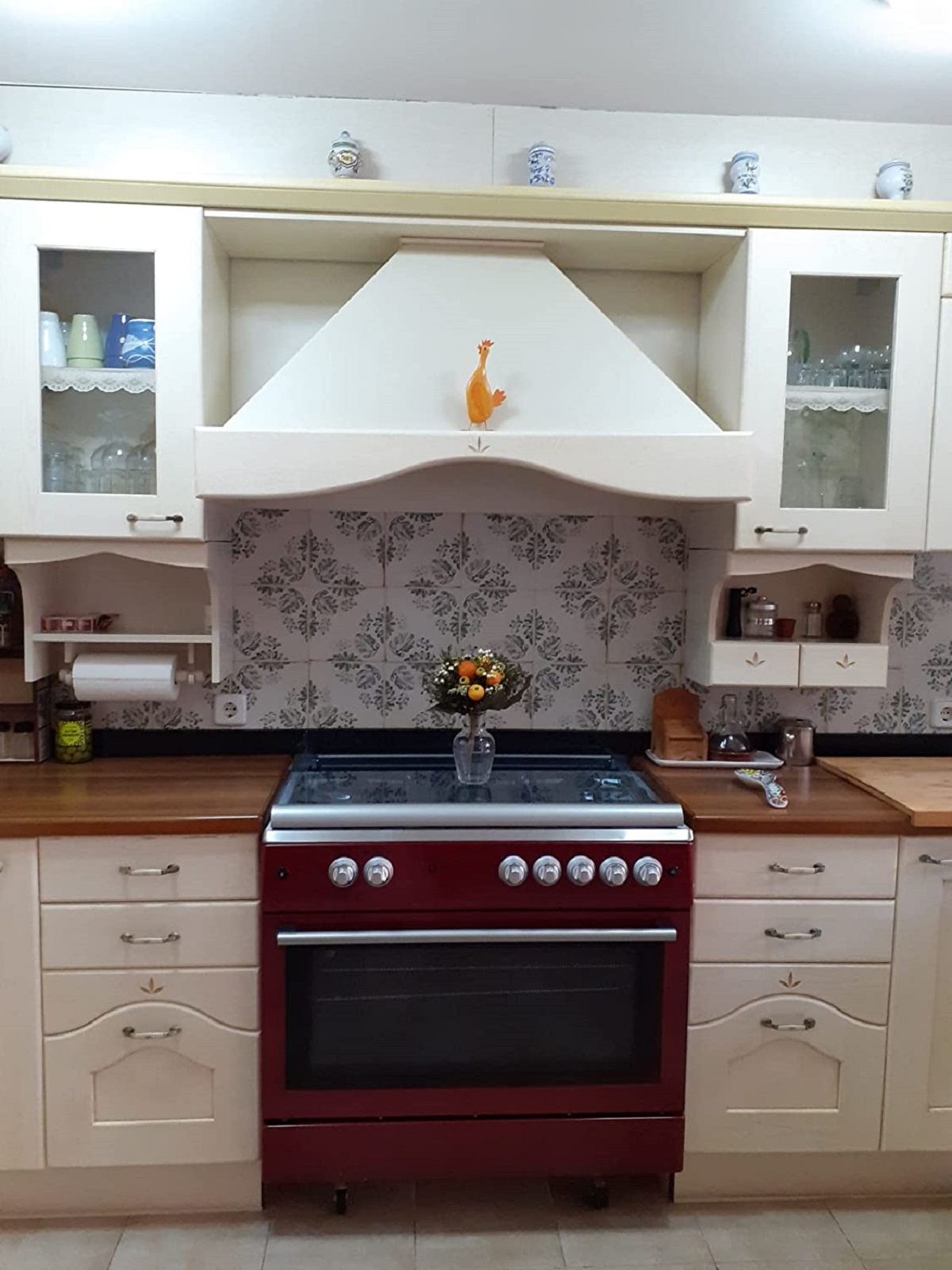 Una cocina con gabinetes color crema, un horno rojo y un salpicadero de azulejos florales.