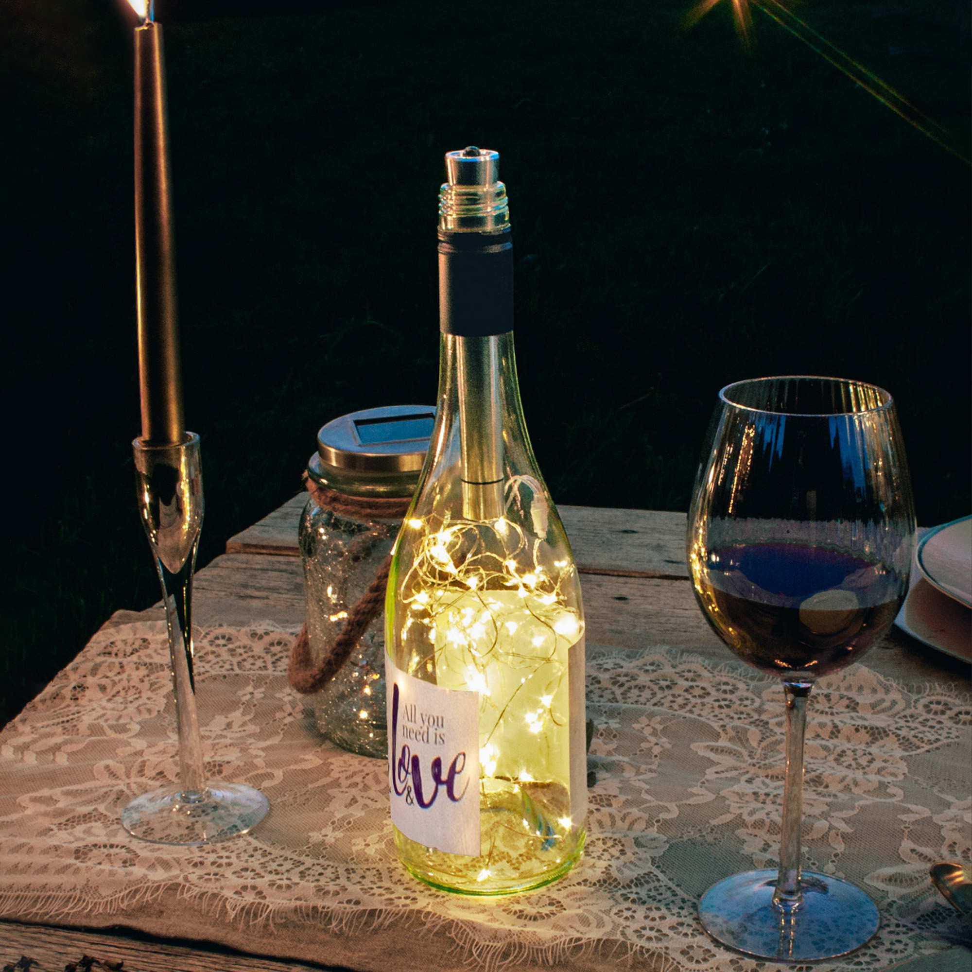 Eine Flasche mit Lichterketten steht auf einem Tisch. Kerze und Weinglas in der Nähe. Kontext: draußen, Nacht.