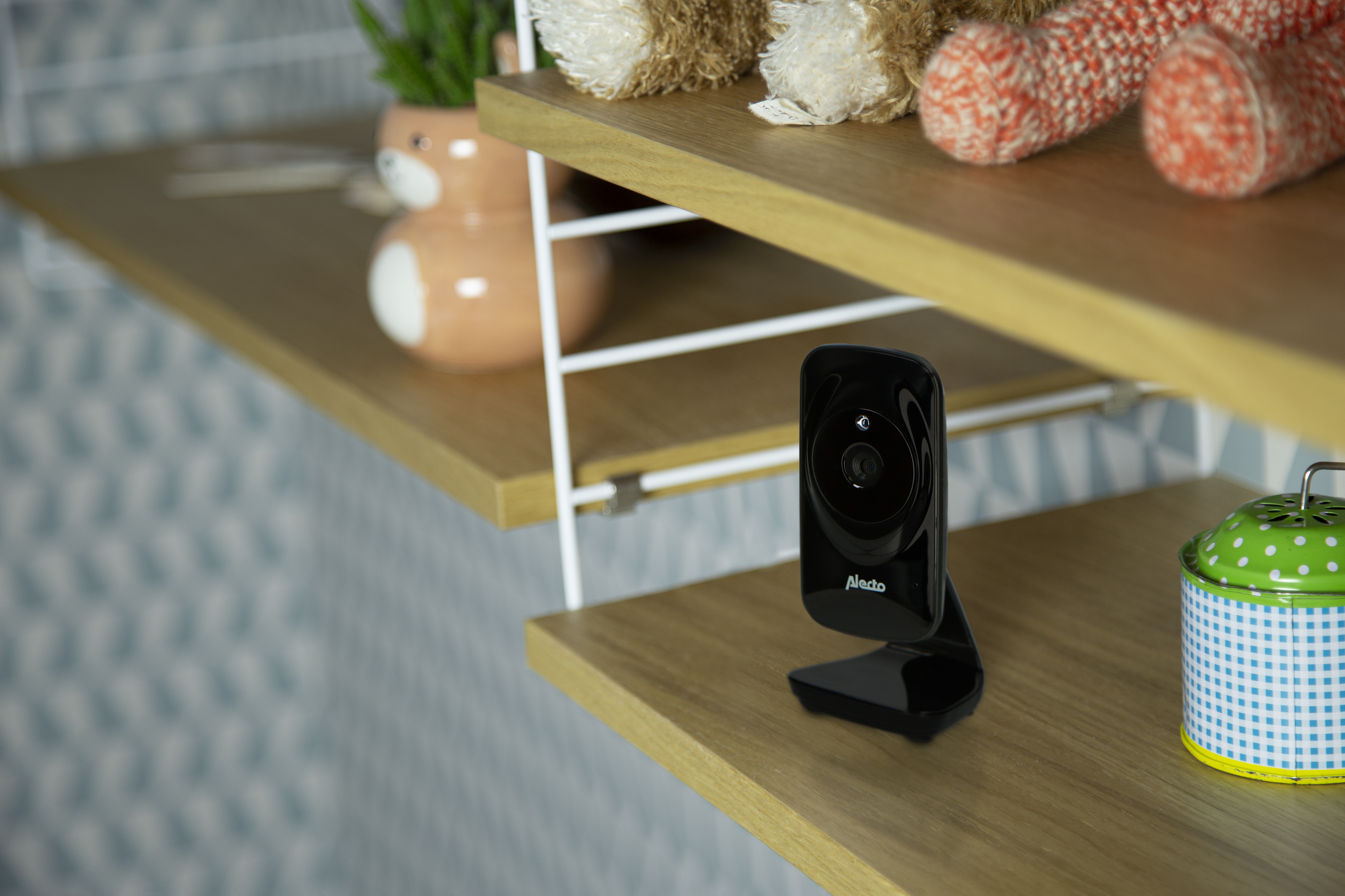 Ein schwarzer Babyphone steht auf einem Holzregal. Weitere Gegenstände befinden sich auf dem Regal, darunter ein Teddybär.