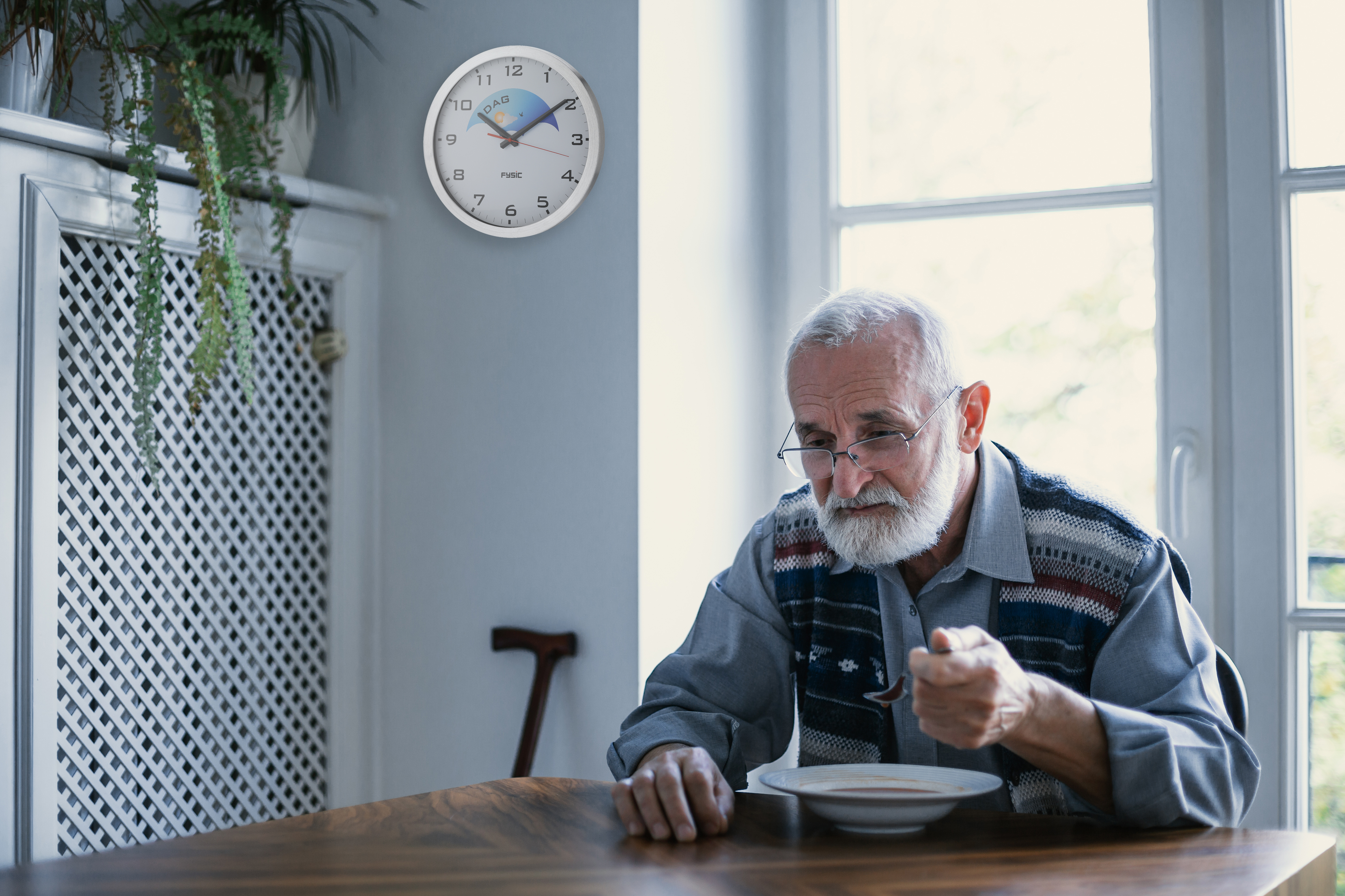 Ein älterer Mann isst Suppe an einem Tisch. Eine Uhr hängt an der Wand hinter ihm.