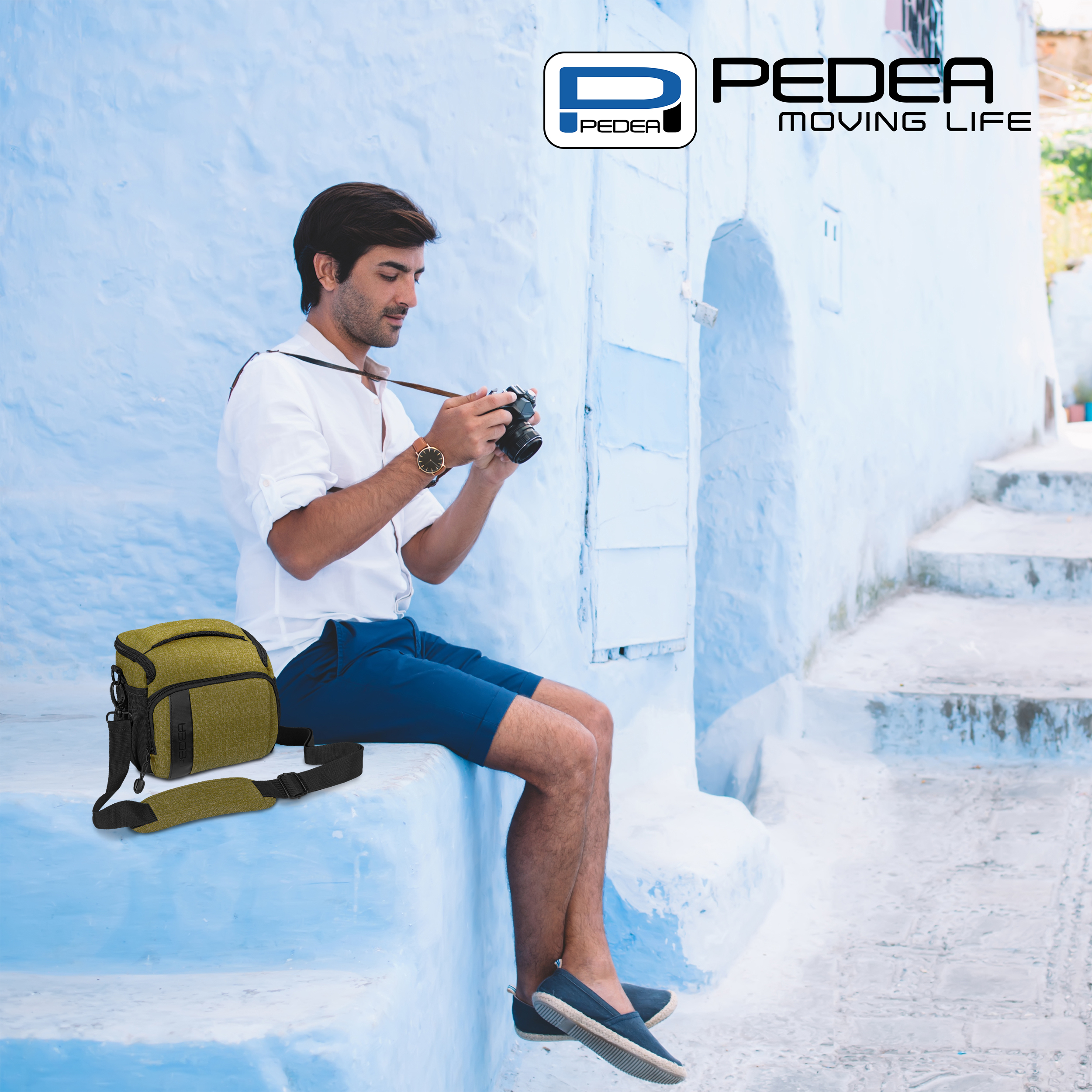 Ein Mann macht ein Foto mit einer Kamera. Er trägt ein weißes Hemd und blaue Shorts. Eine grüne Tasche ist dabei.