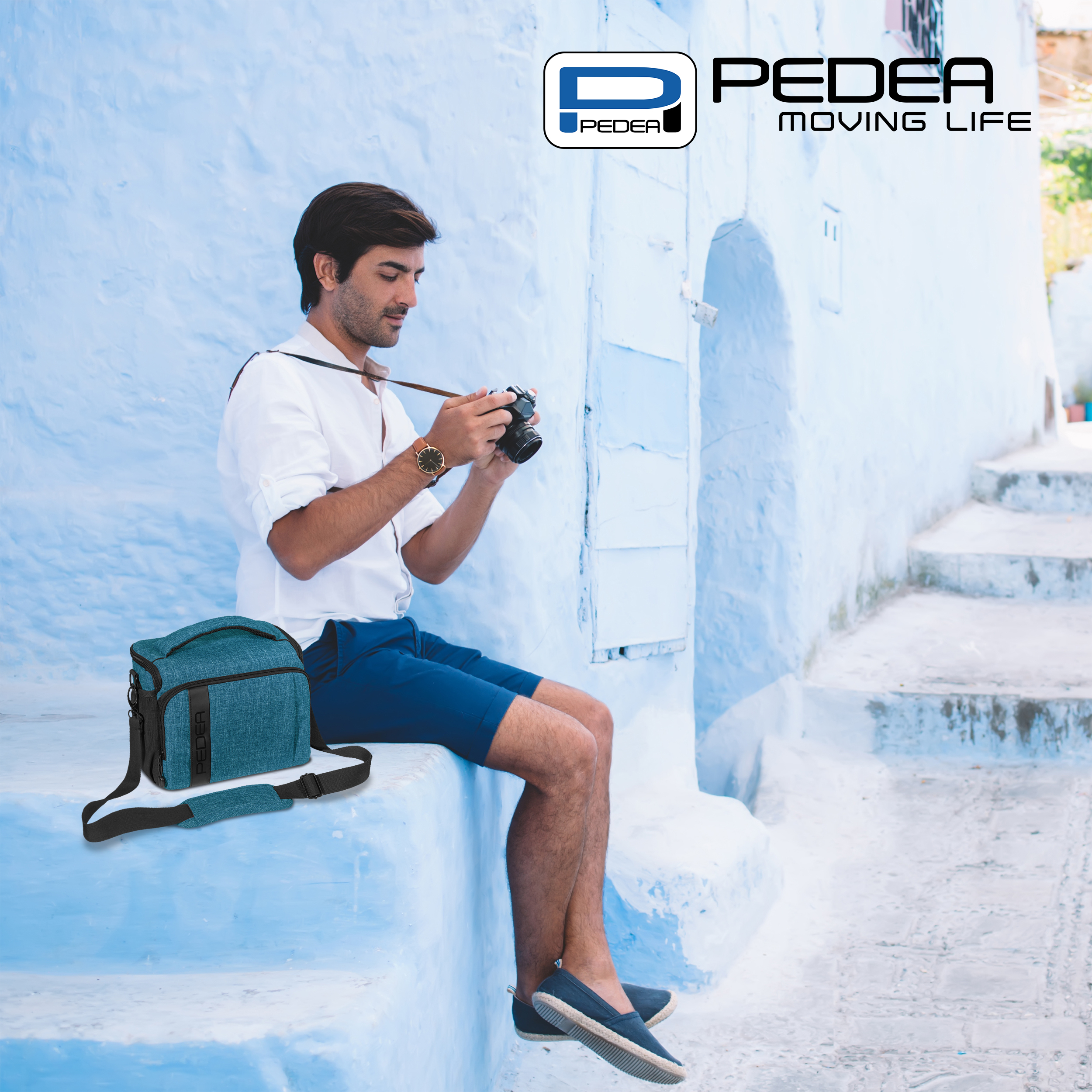 Ein Mann in weißem Hemd und blauen Shorts macht ein Foto mit einer Kamera. Eine blaue Tasche liegt neben ihm an einer Wand.