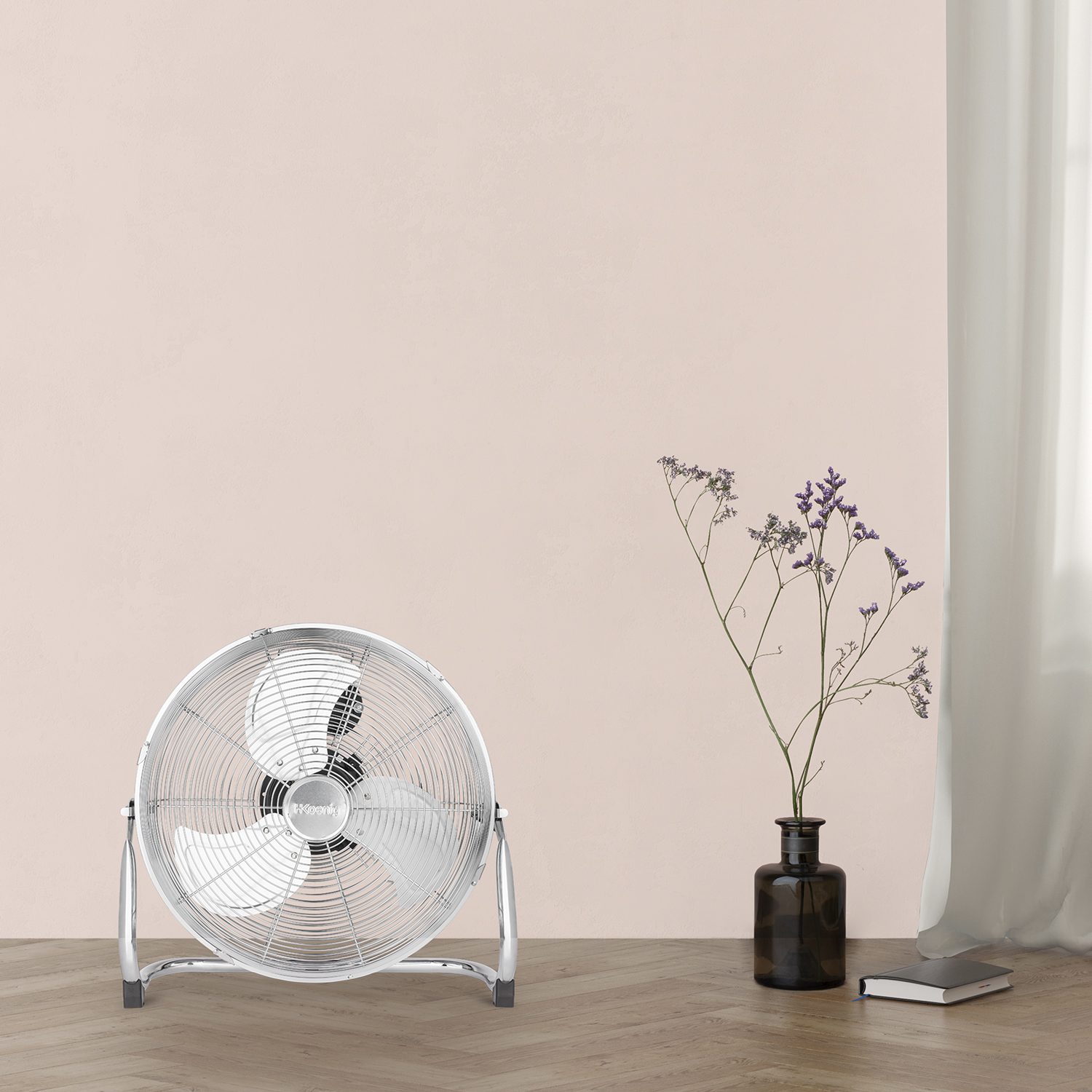 Un ventilatore argentato su un pavimento in legno, accanto a un vaso con fiori e un libro.