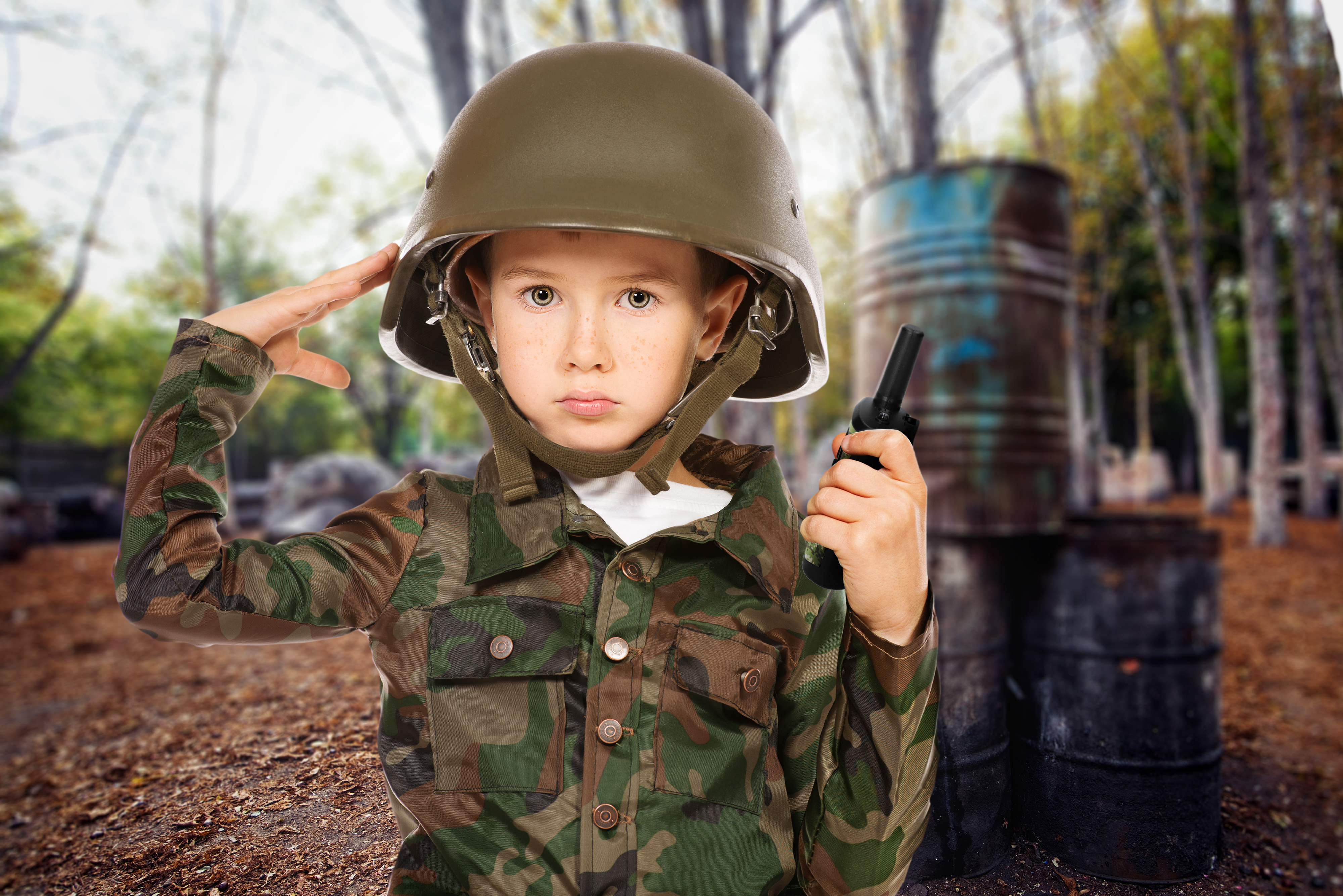 Młody chłopiec w kamuflażu i hełmie salutuje. Trzyma krótkofalówkę, w tle las.