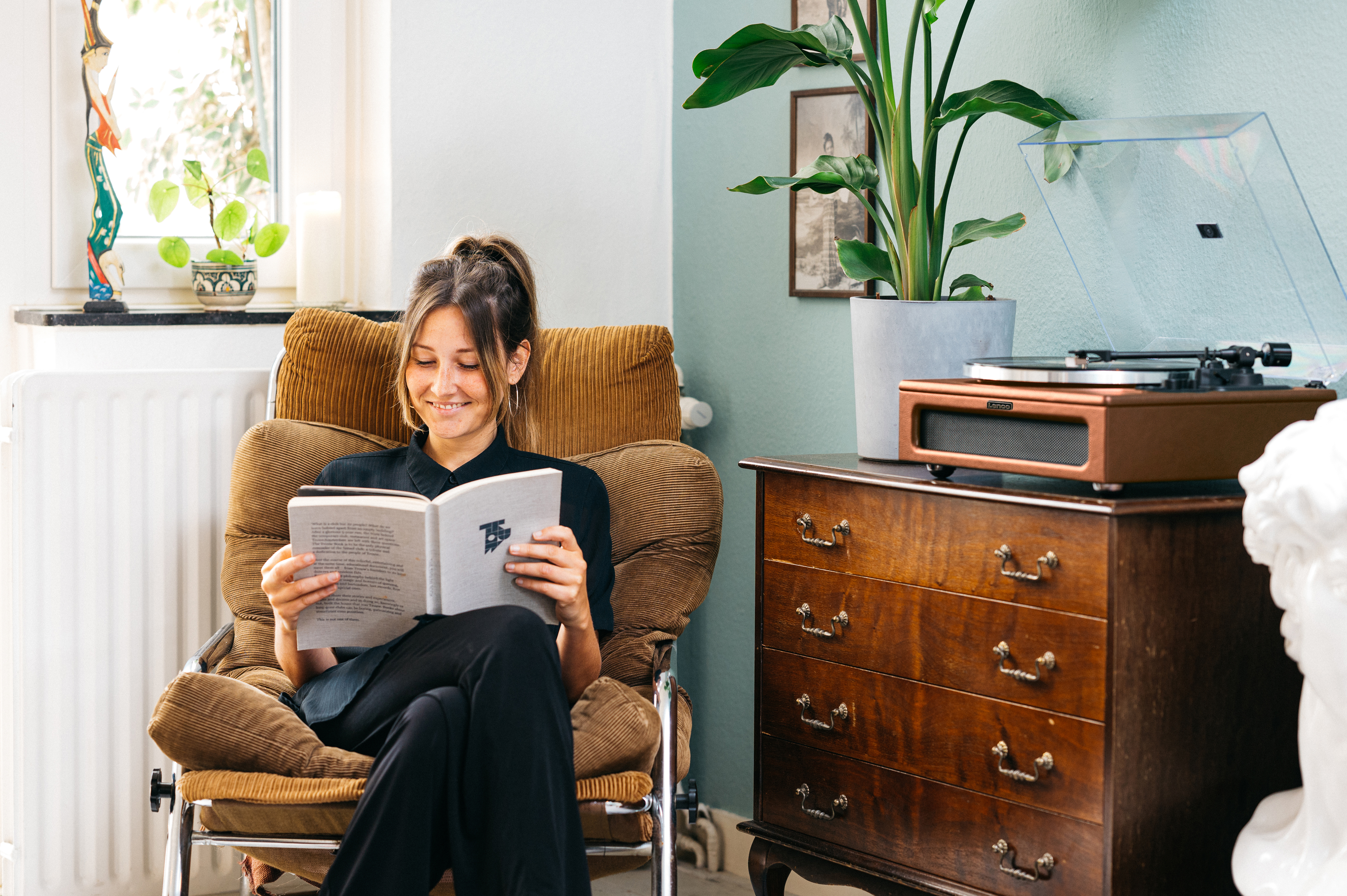 Kobieta siedzi na krześle czytając książkę w pobliżu gramofonu.