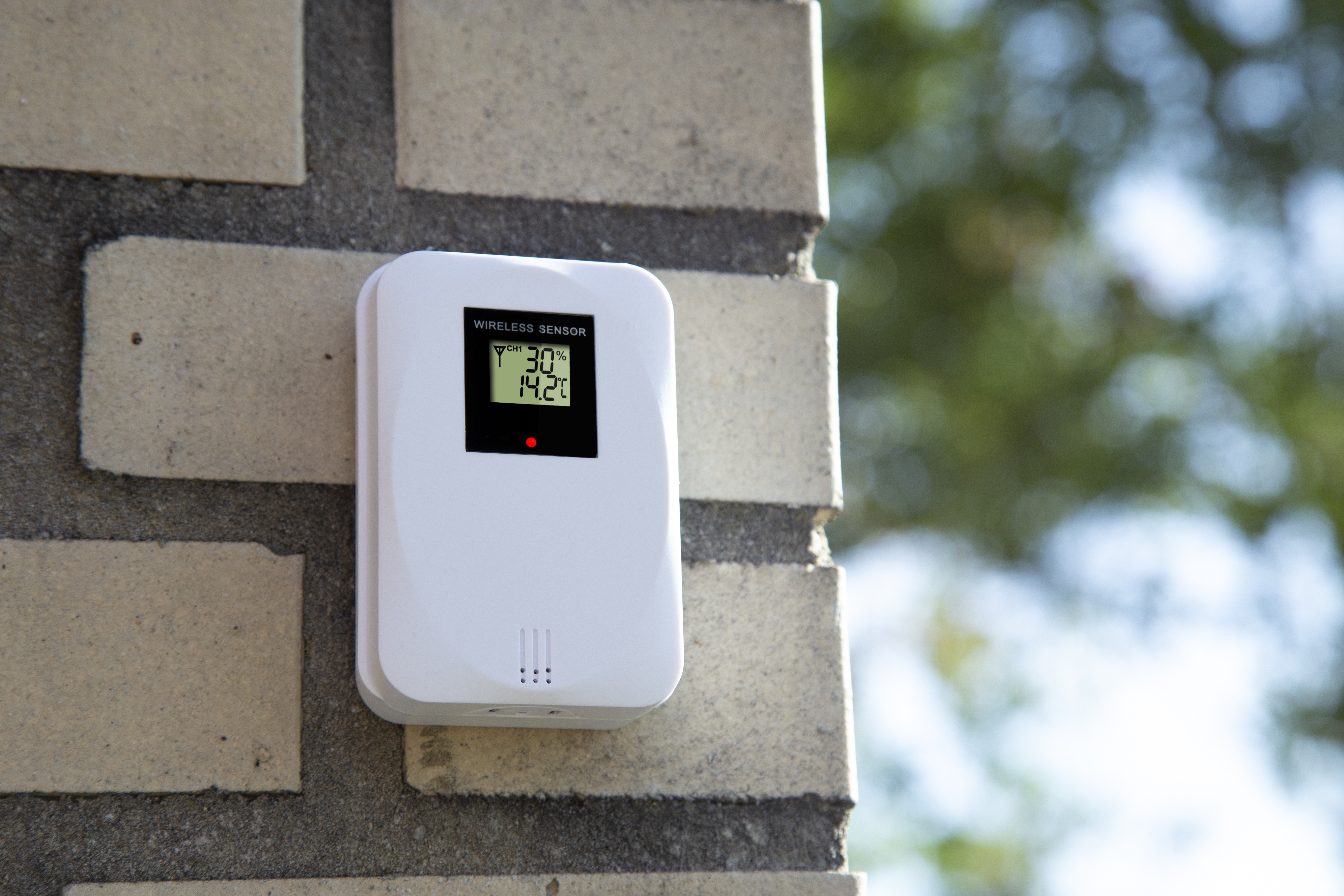 Biały zewnętrzny czujnik pogody zamontowany na ceglanej ścianie. Wyświetla odczyty temperatury i wilgotności.