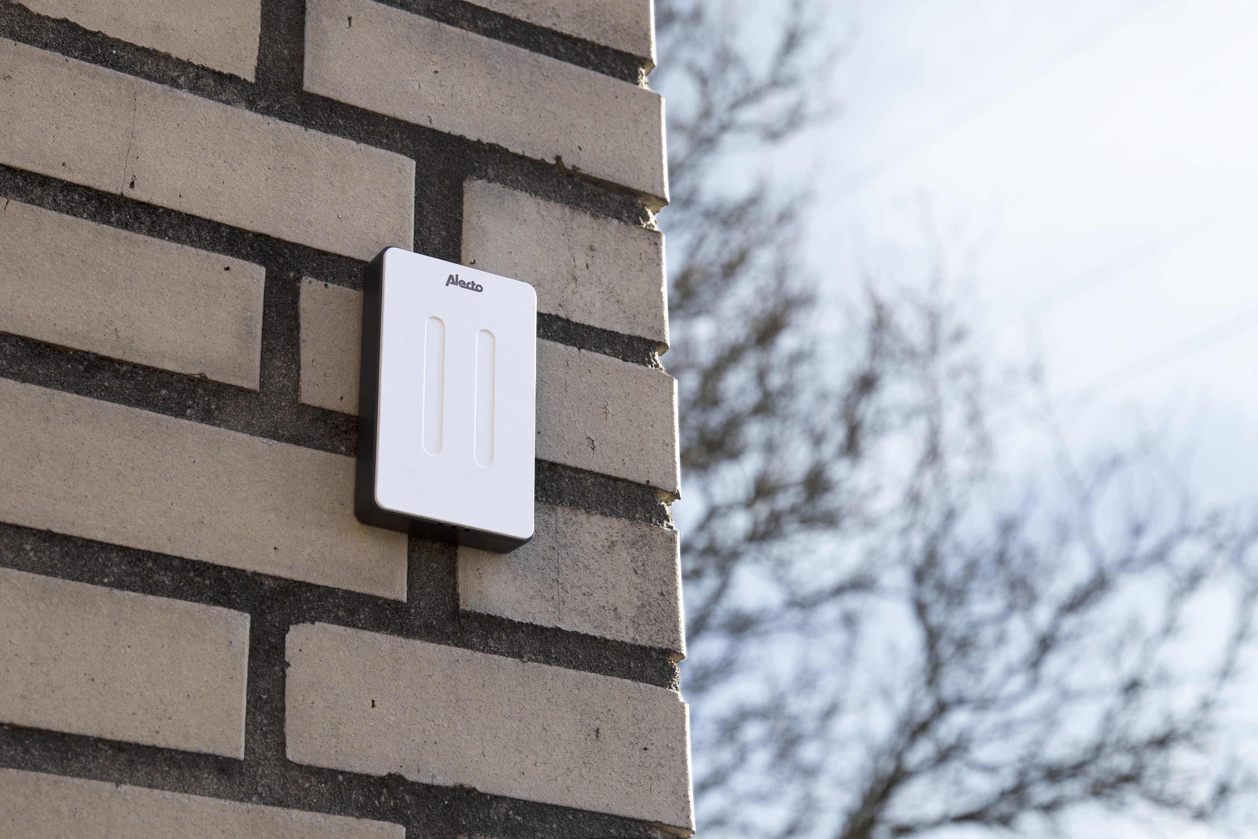 A white smart home device is mounted on a brick wall. The sky and tree branches are visible in the background.