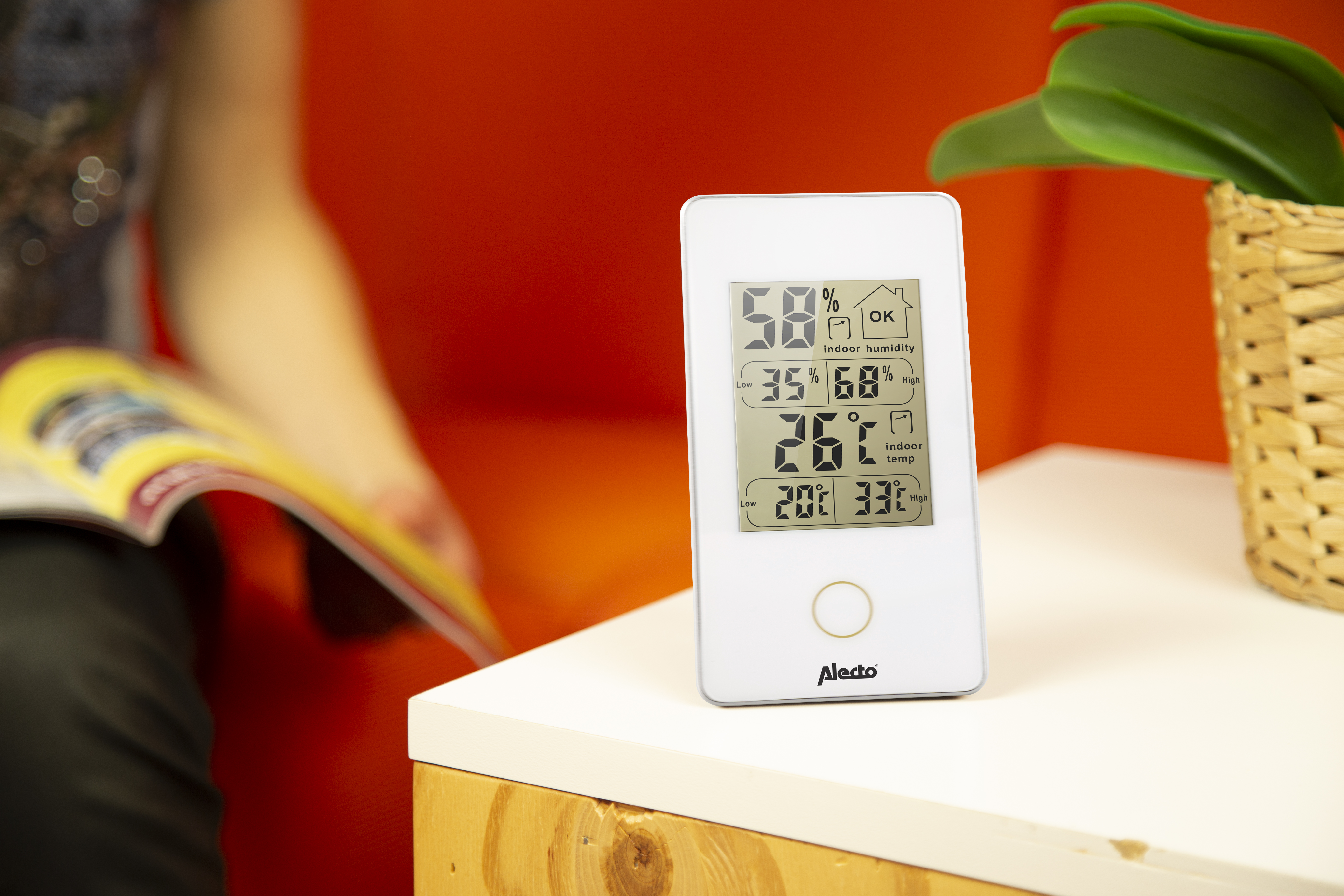 Termometro digitale bianco che mostra temperatura e umidità interna, con una persona sullo sfondo.