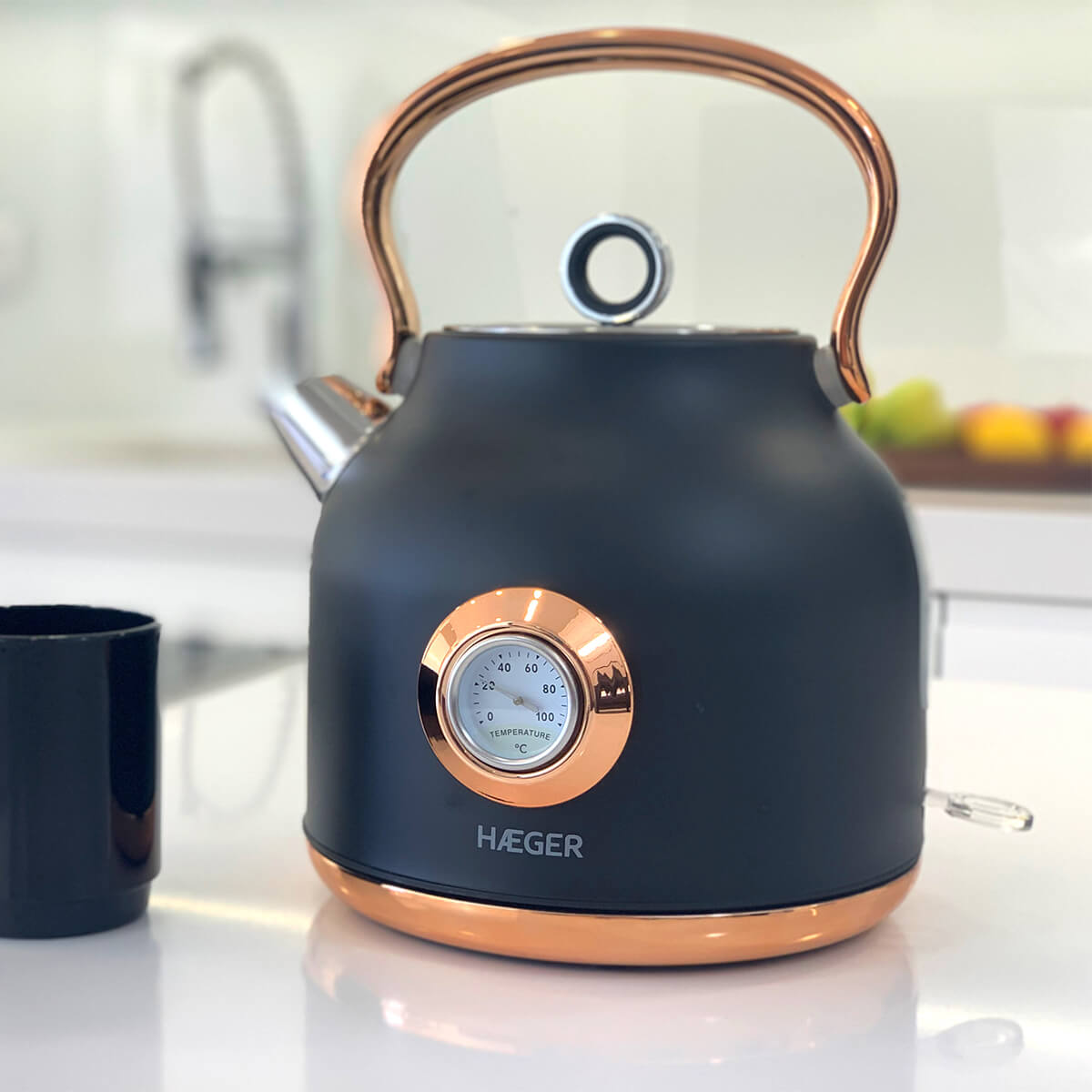 Bollitore nero e rame con indicatore di temperatura, su un piano bianco. Sfondo cucina, con una tazza nera a sinistra.