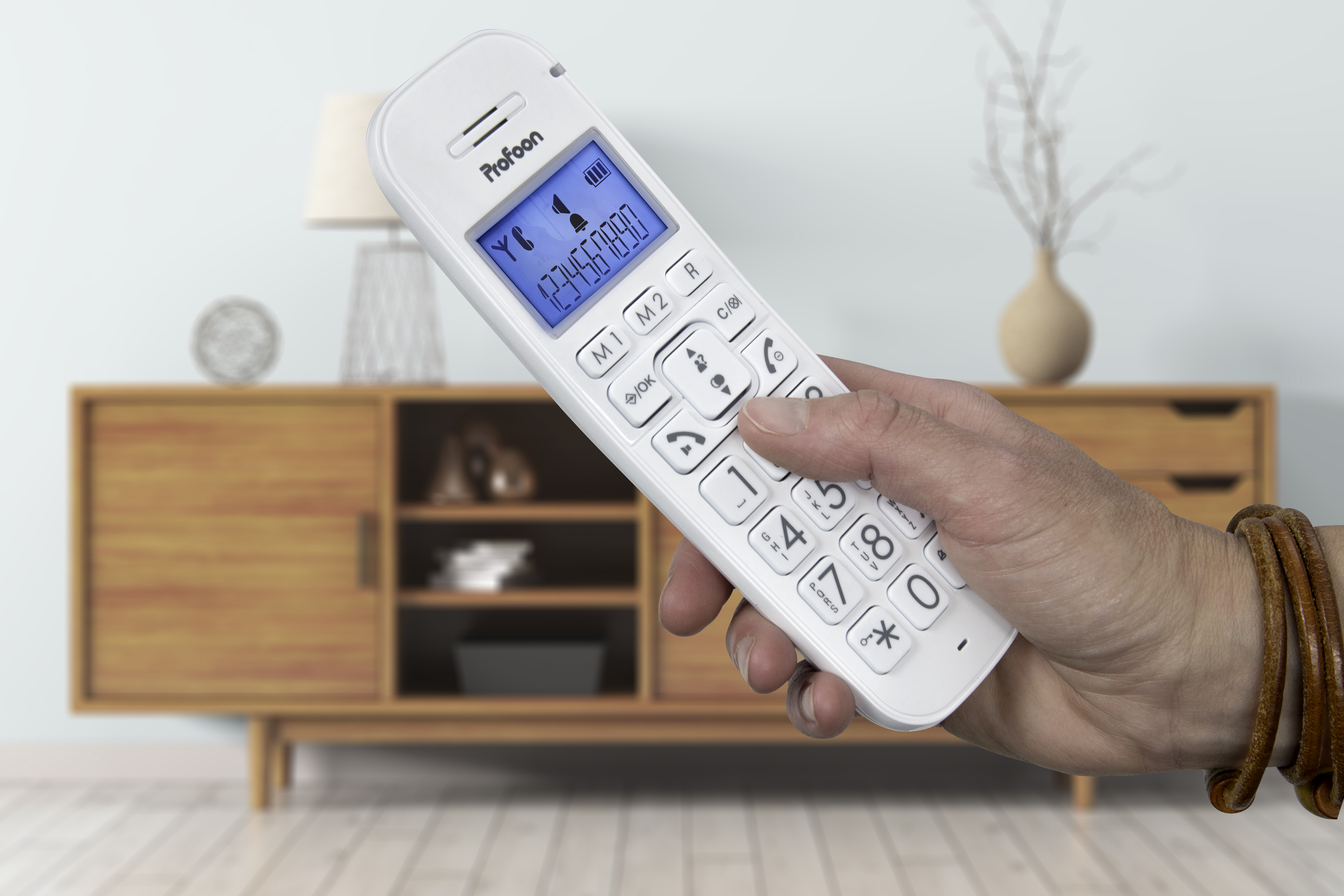 Eine Hand, die ein weißes schnurloses Telefon hält. Ein Holzschrank und Dekoration befinden sich im Hintergrund.