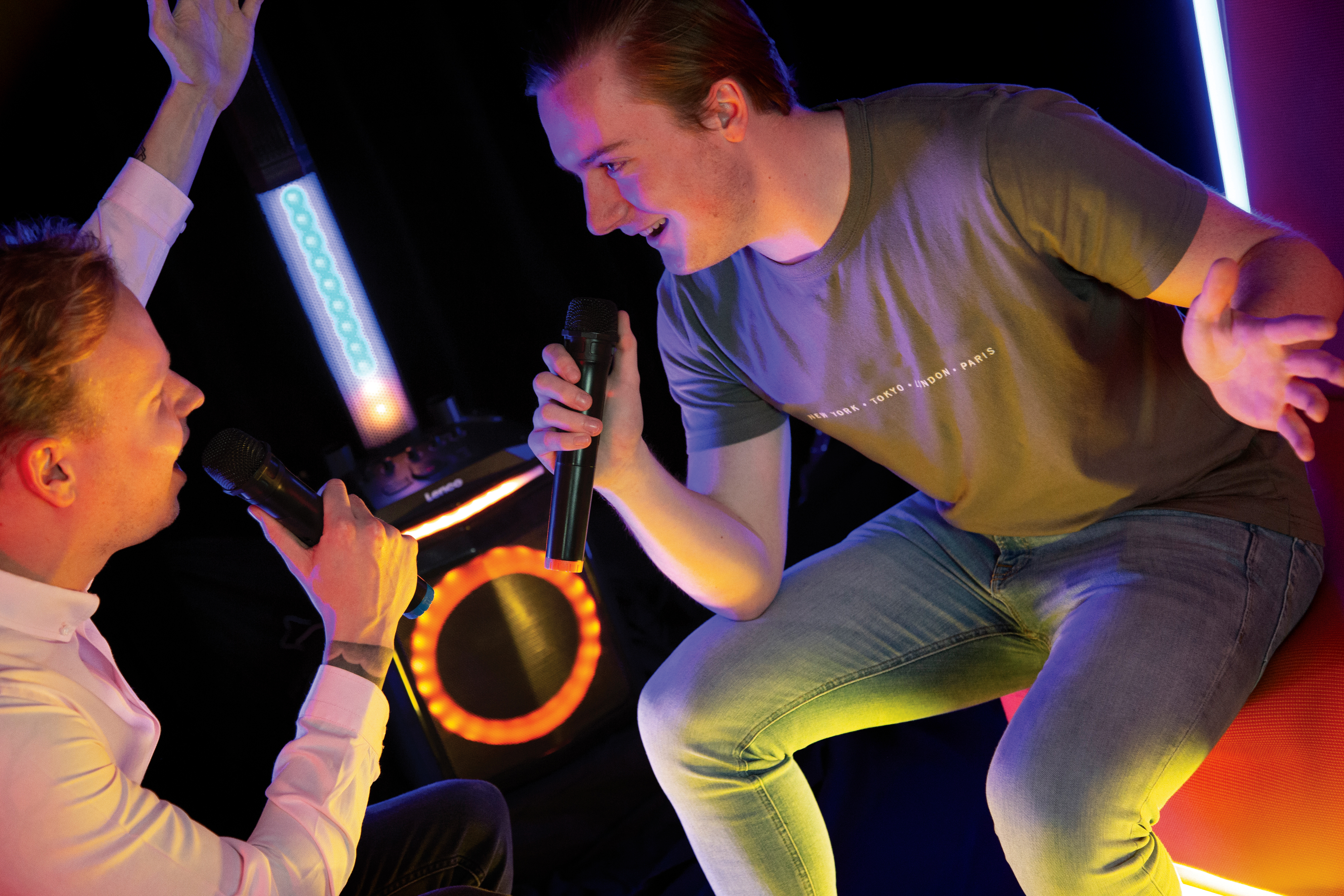 Dwóch mężczyzn śpiewa do mikrofonów, jeden podnosi rękę, przed głośnikiem ze światłami. Ciemne tło.