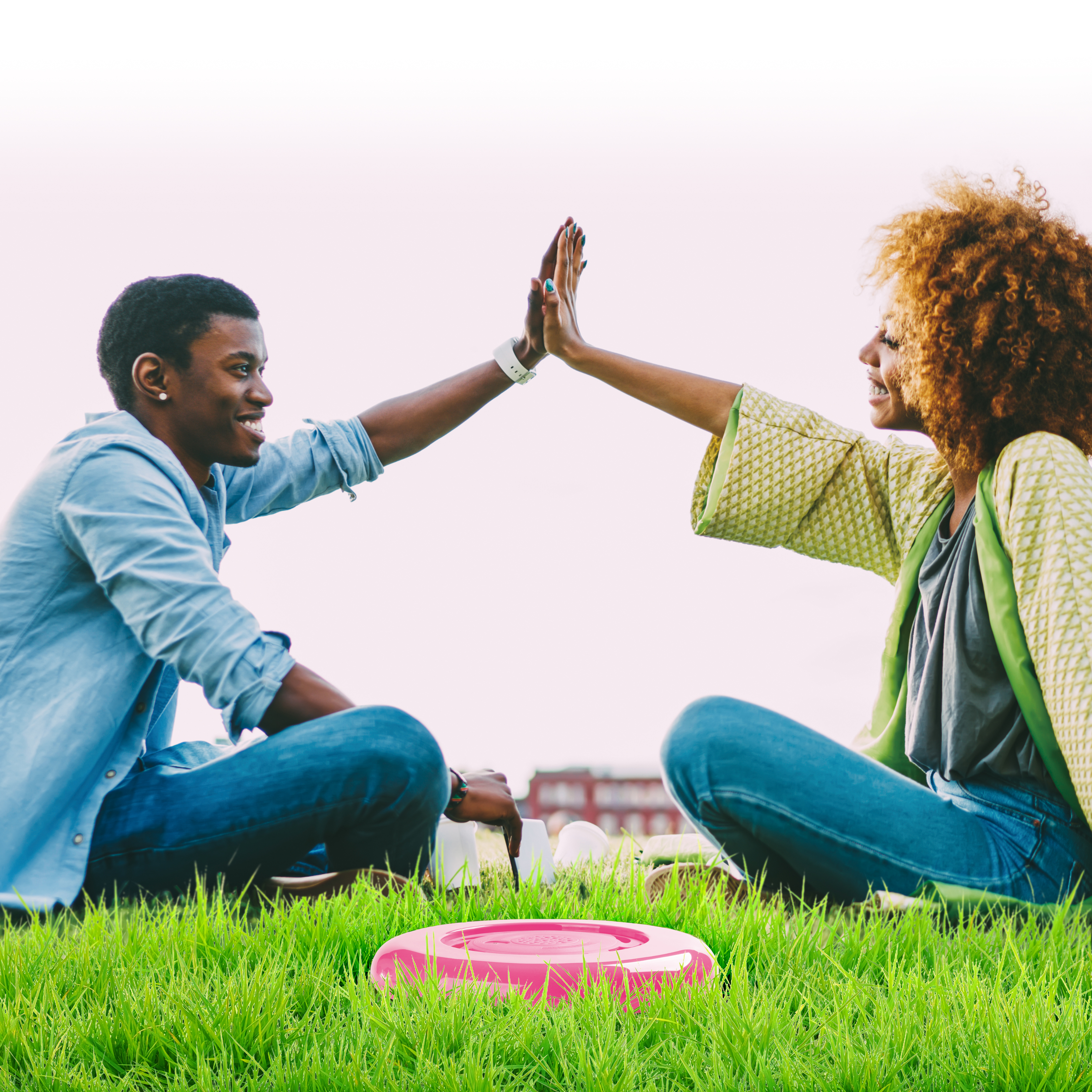 Twee mensen geven high-five, zitten op gras, roze frisbee vooraan. Heldere hemel op de achtergrond.