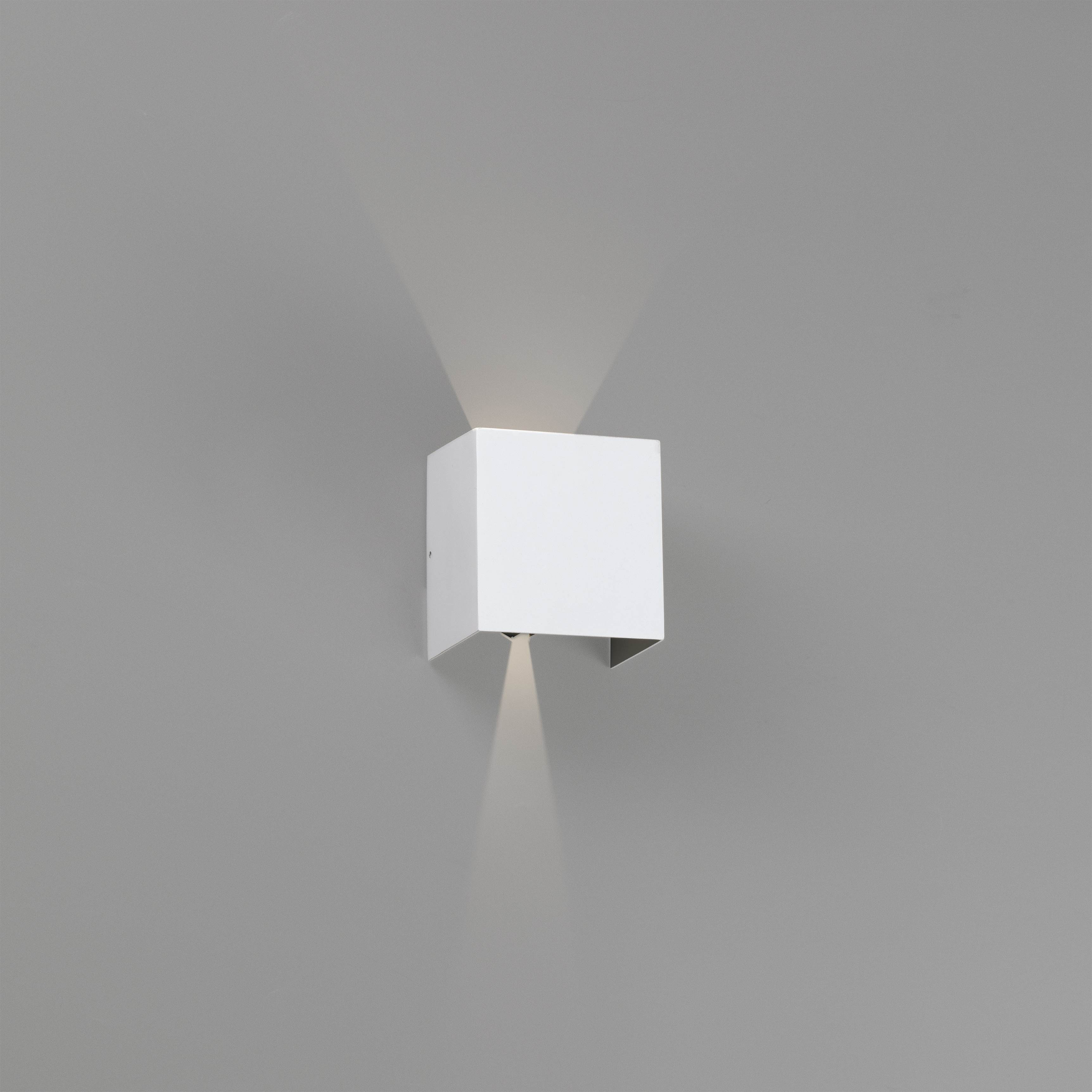 Lampada da parete bianca a forma di cubo. Ha una luce rivolta verso il basso. È montata su un muro grigio.