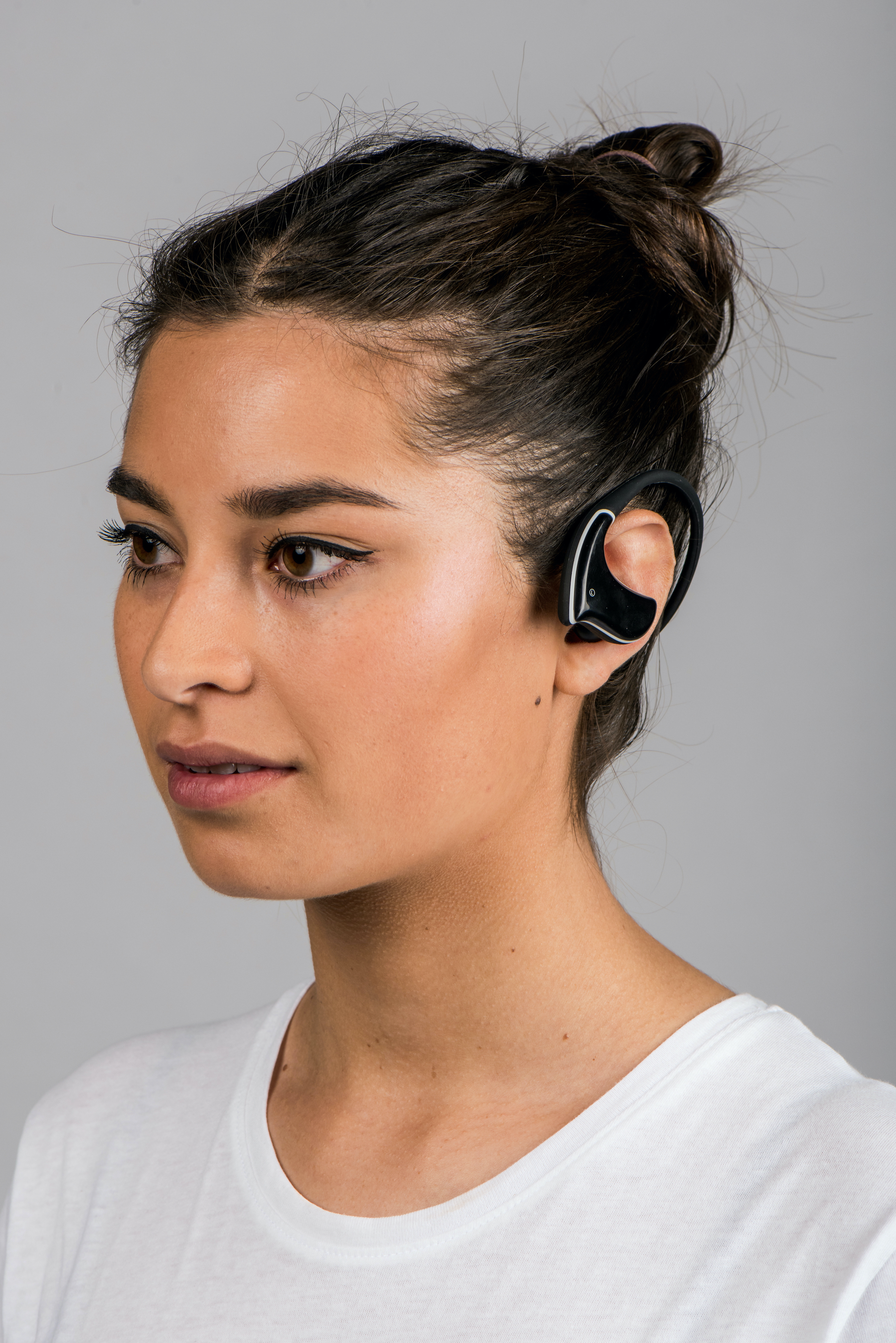 Una donna con un auricolare nero, vista di profilo, con una maglietta bianca, su sfondo grigio.