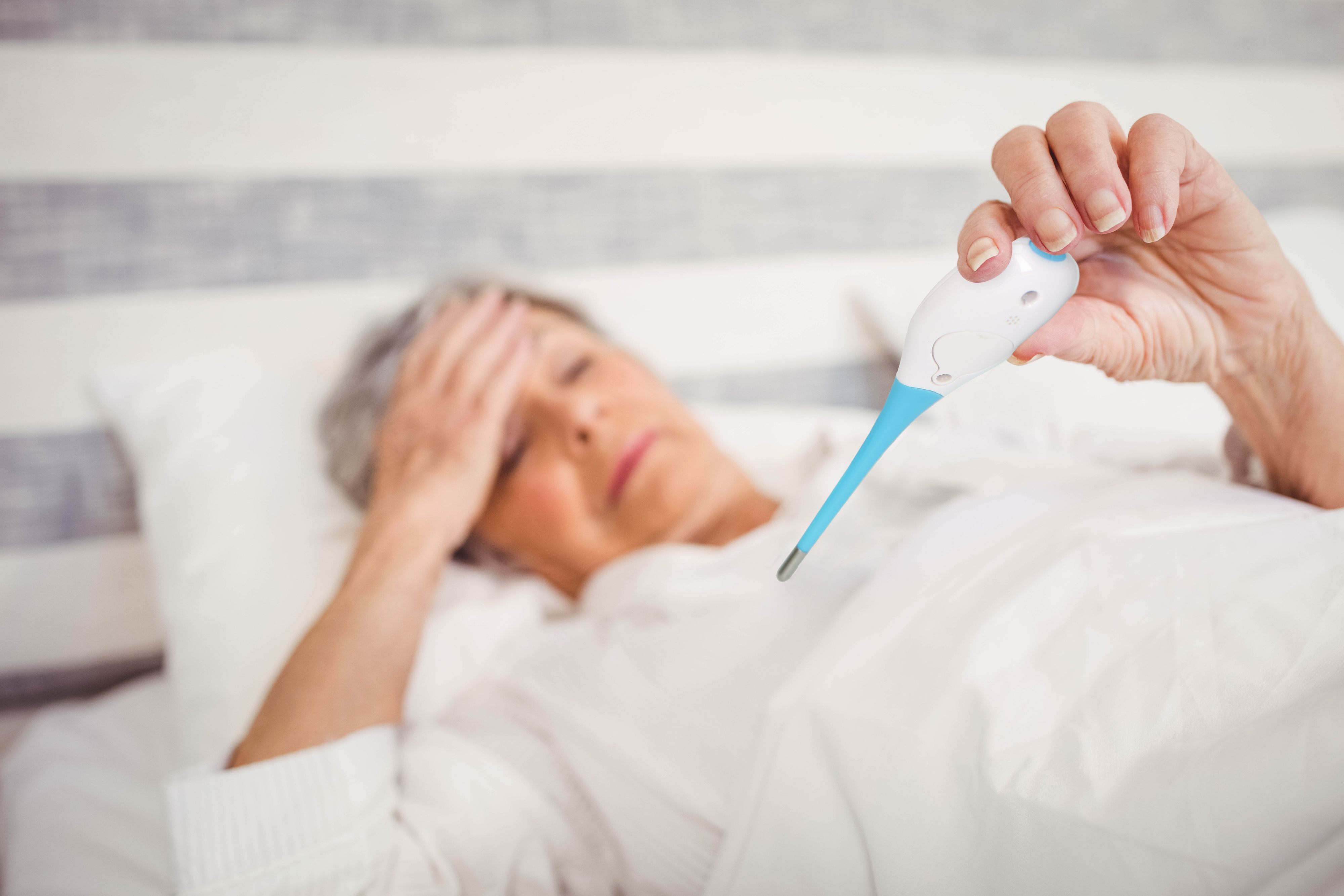 Eine ältere Frau im Bett hält ein Thermometer und berührt ihre Stirn. Sie sieht unwohl aus.