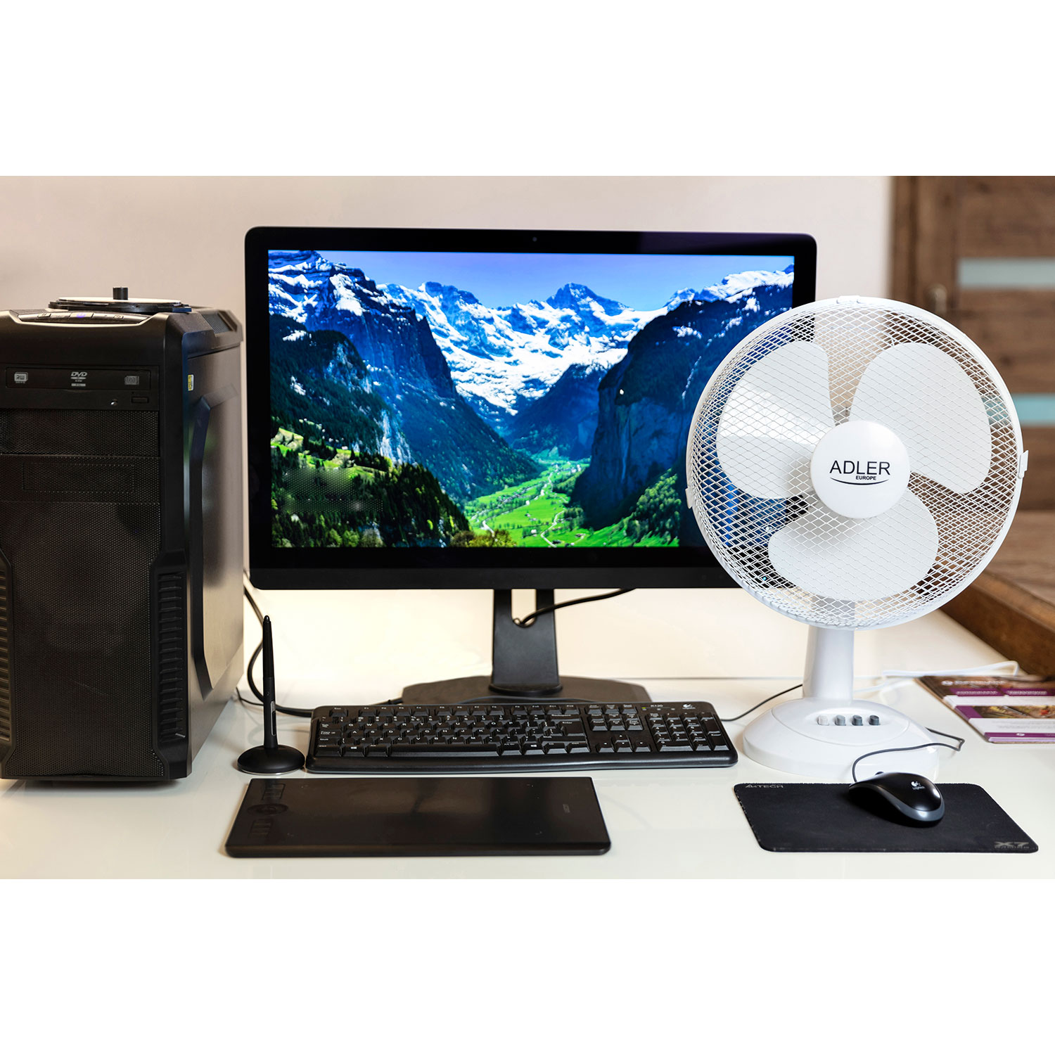 Un escritorio con computadora, monitor, teclado, ratón, ventilador y tableta de dibujo.