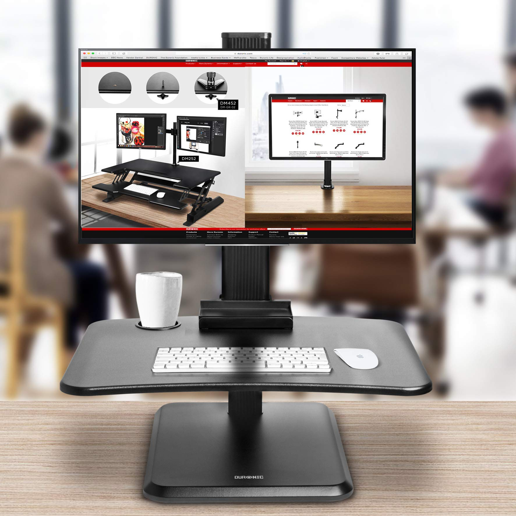 Un soporte de monitor con un monitor, teclado, ratón y una taza, con personas borrosas en el fondo.
