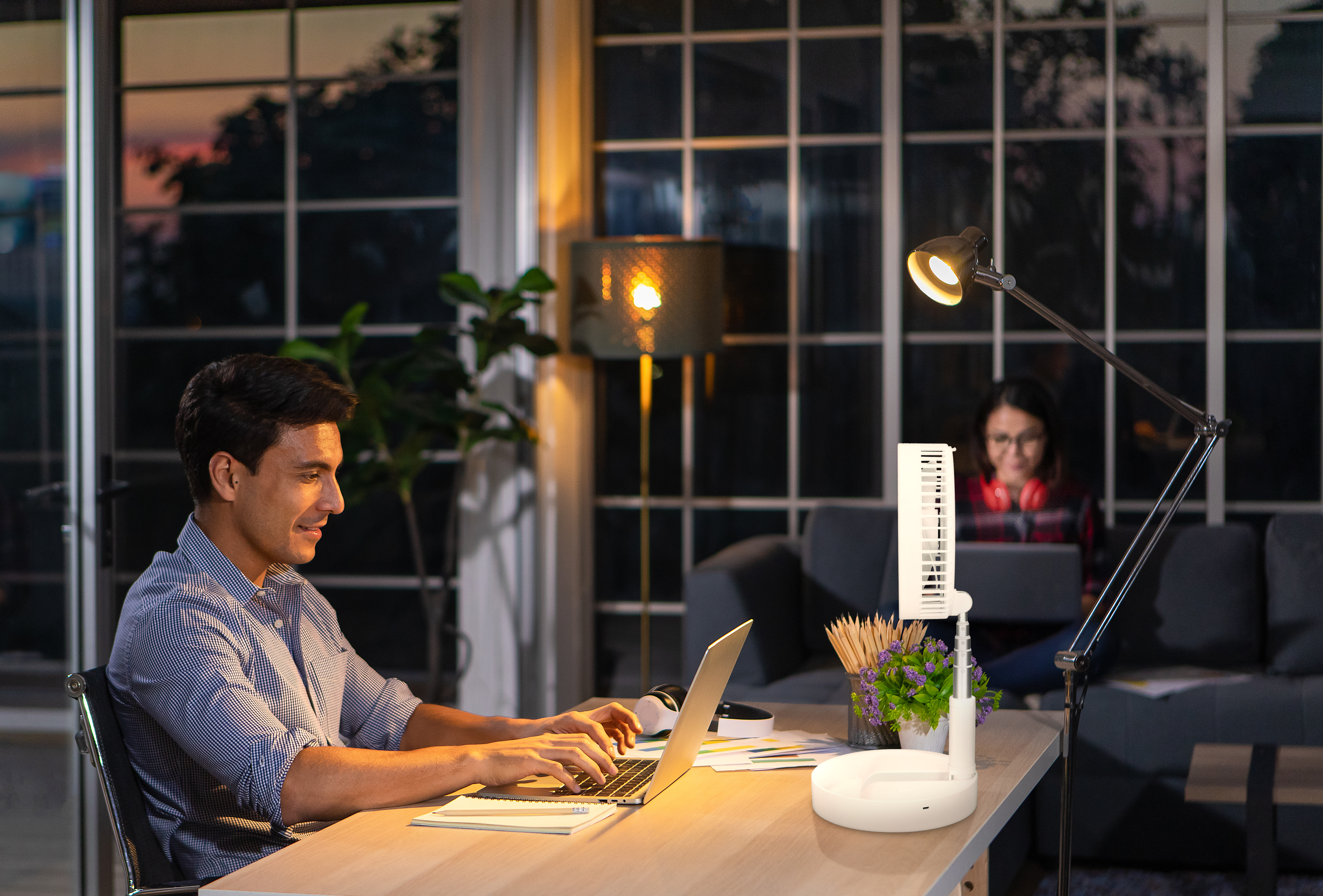 Mann arbeitet am Laptop, Frau im Hintergrund. Schreibtischventilator und -lampe sind sichtbar.