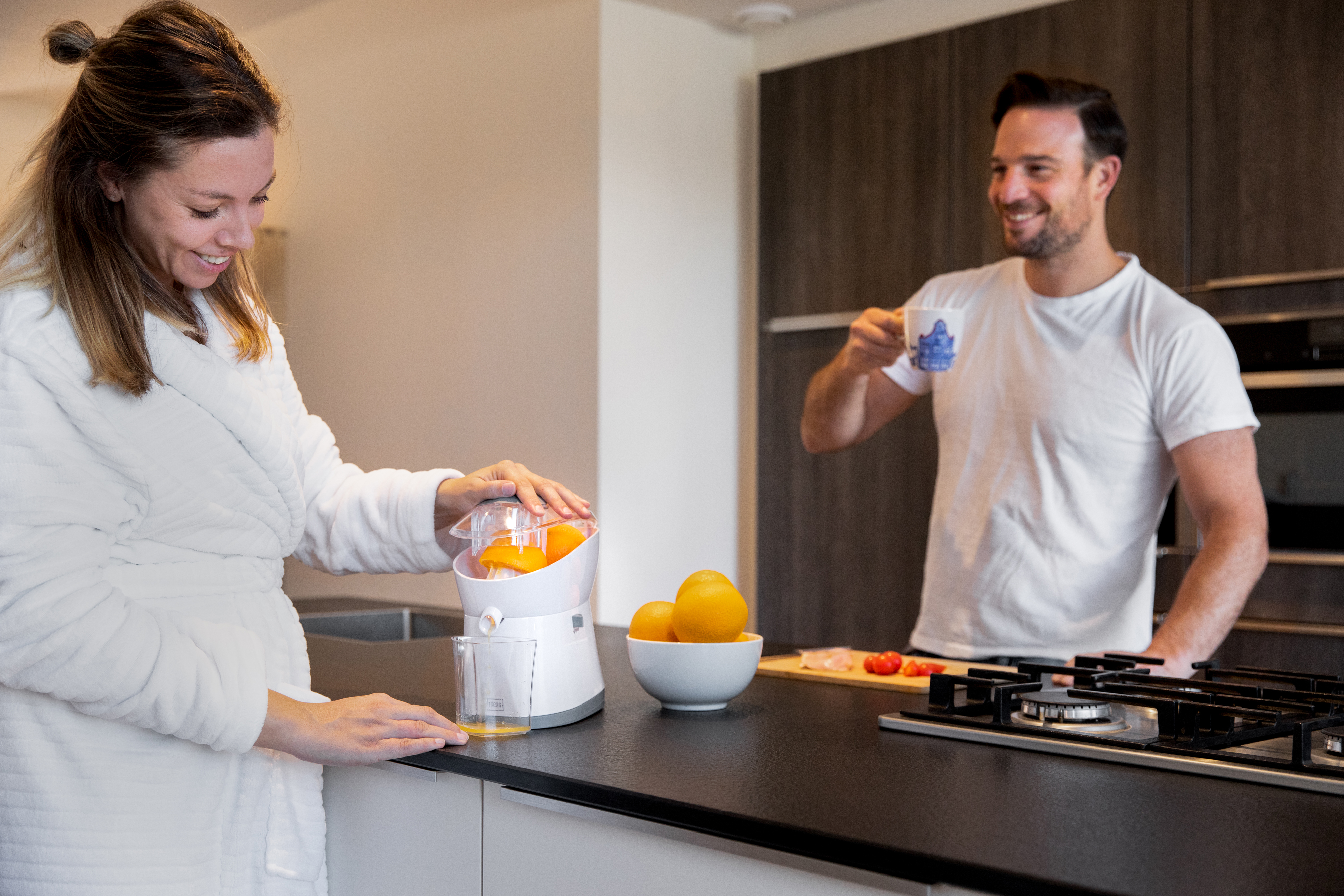 Frau im Bademantel presst Orangen in der Küche. Mann im Hintergrund hält eine Tasse.