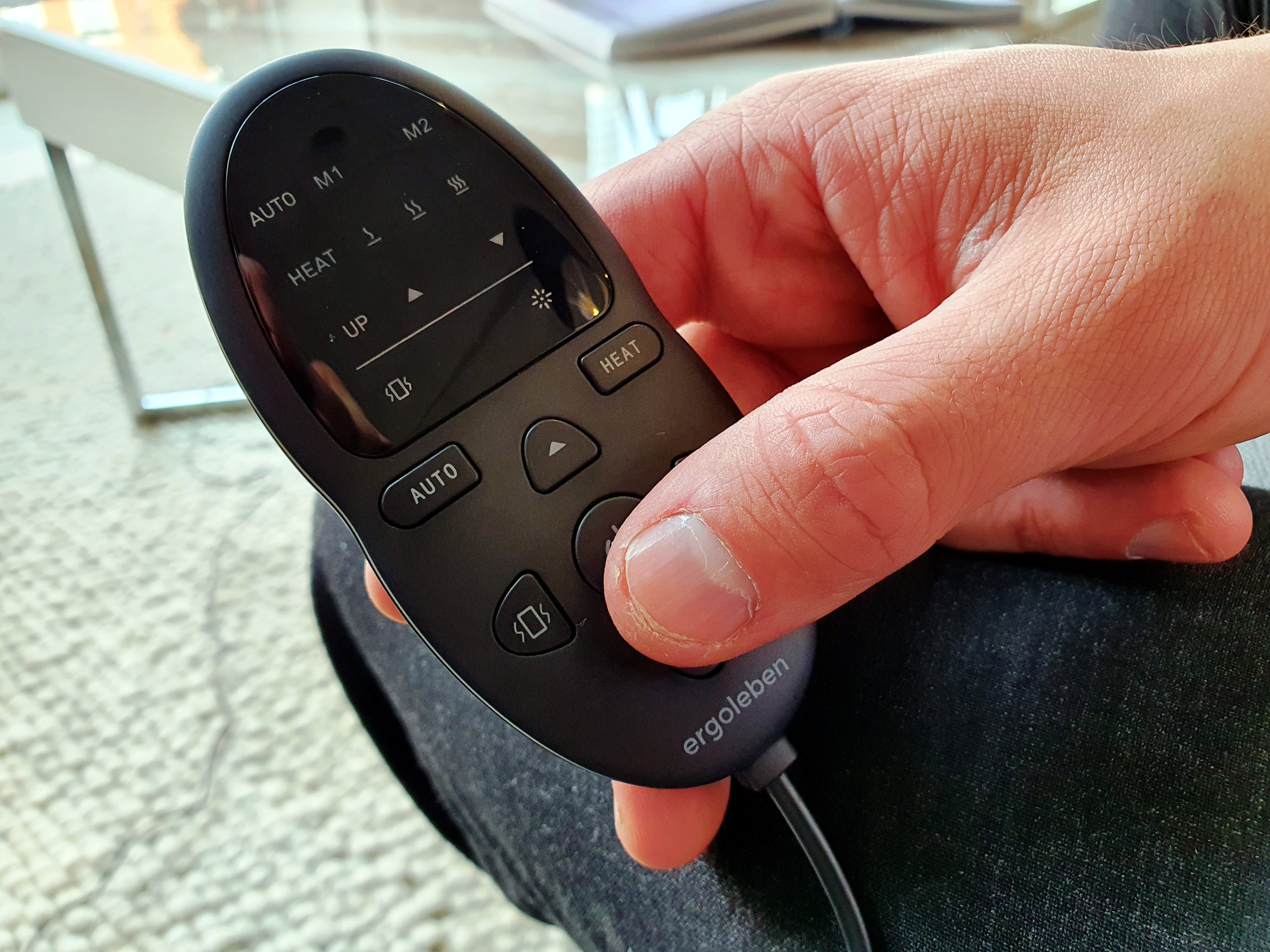 Eine Hand, die eine schwarze Fernbedienung mit den Tasten 'AUTO', 'HEAT' und anderen Bedienelementen hält. Der Hintergrund ist unscharf.