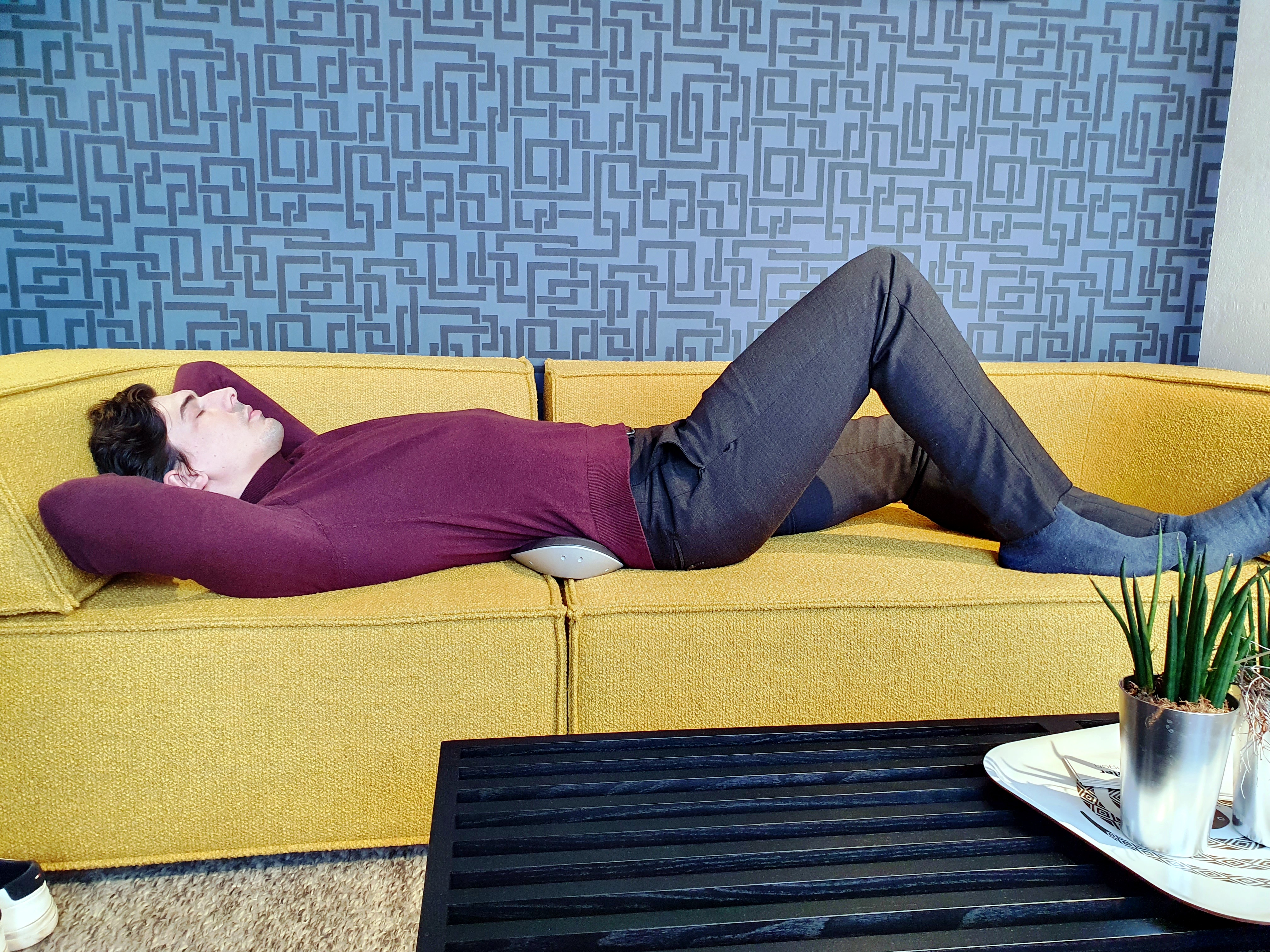 Mann liegt auf gelbem Sofa, Lendenstütze am Rücken, Tisch davor.
