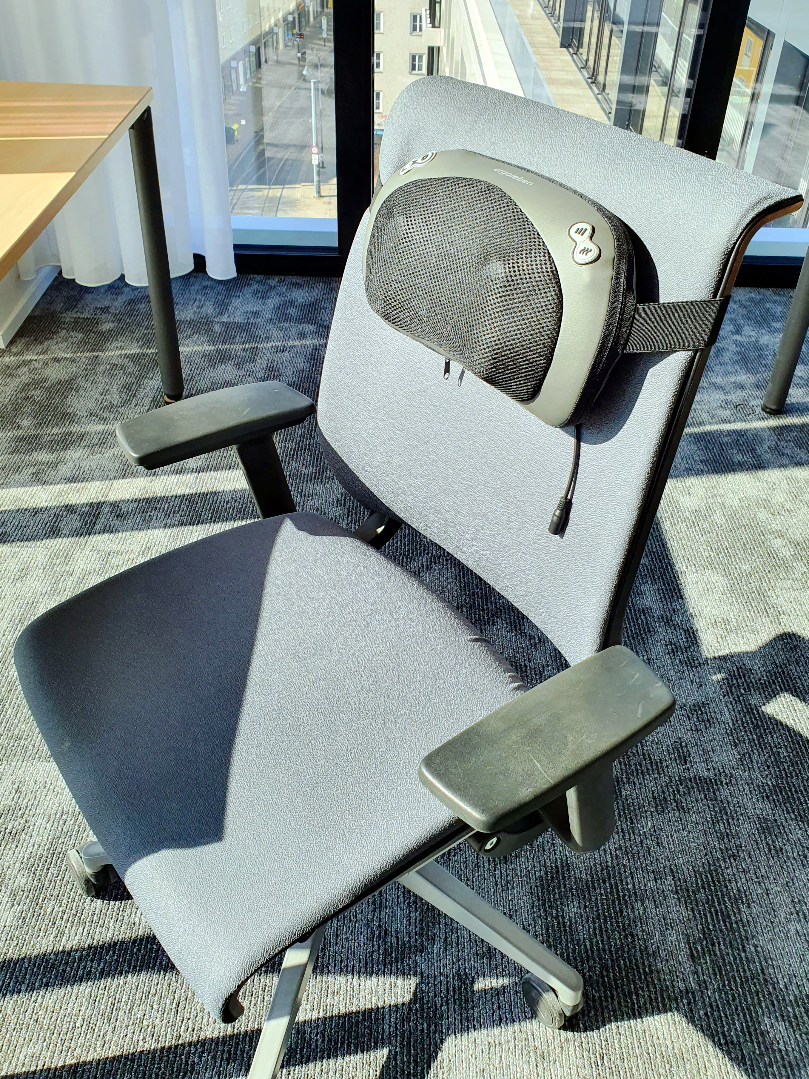 Grauer Bürostuhl mit einem schwarzen Rückenmassagegerät. Der Stuhl befindet sich in einem Büroumfeld.