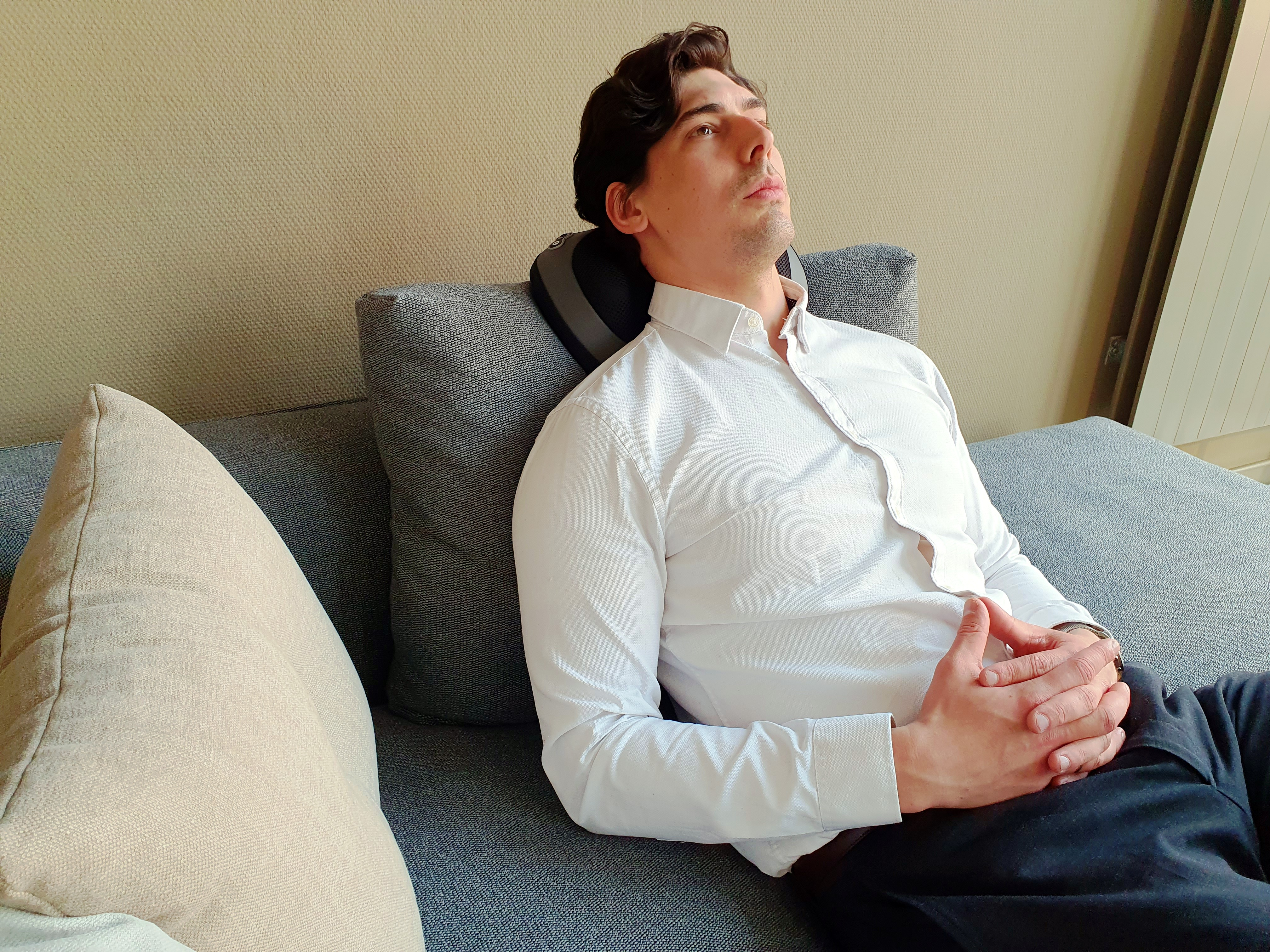 Ein Mann auf einem grauen Sofa mit weißem Hemd und Nackenmassagegerät. Er schaut nach oben.