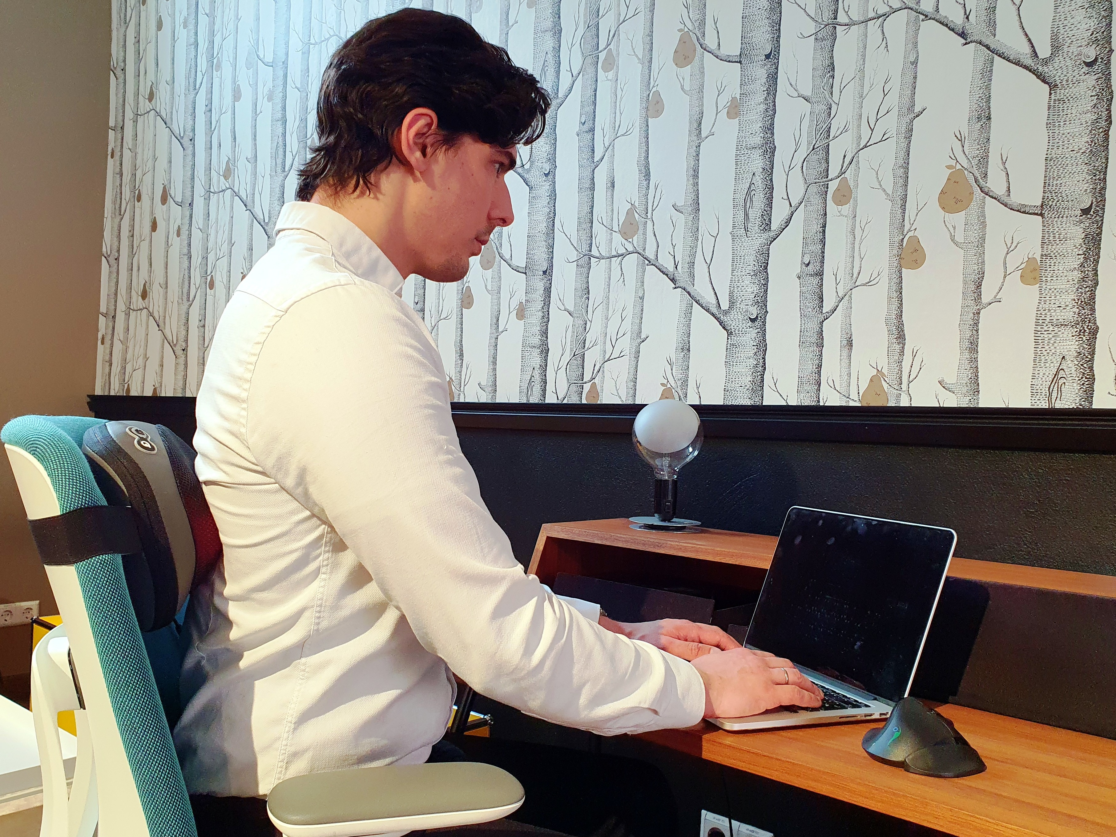 Ein Mann arbeitet an einem Laptop an einem Holztisch. Es gibt eine Lampe, einen Stuhl und eine gemusterte Wand.