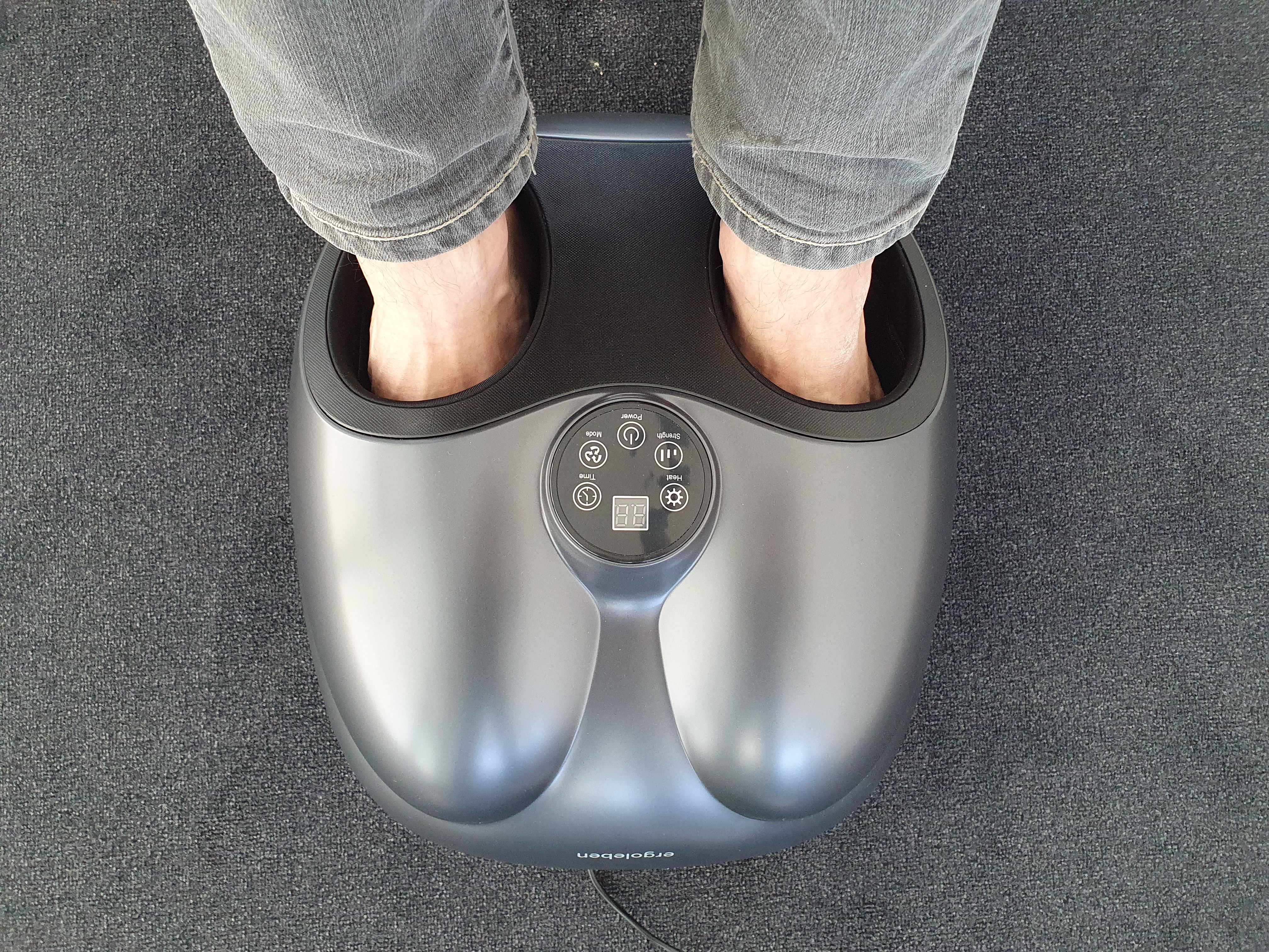 Füße in einem Fußmassagegerät. Graue Füße, schwarzes Massagegerät und grauer Teppich.