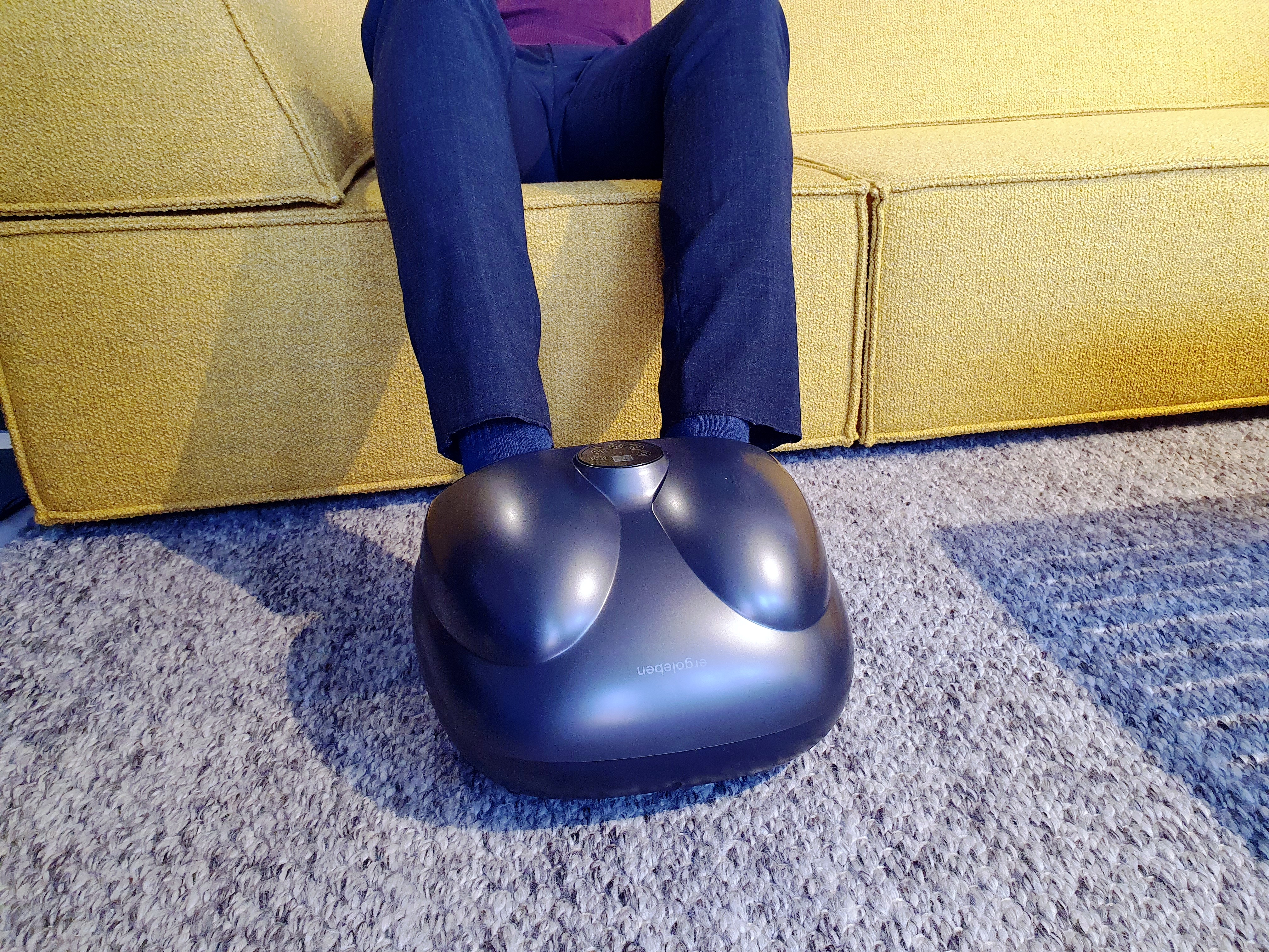 Eine Person sitzt auf einem gelben Sofa, die Füße auf einem schwarzen Fußmassagegerät auf einem grauen Teppich.
